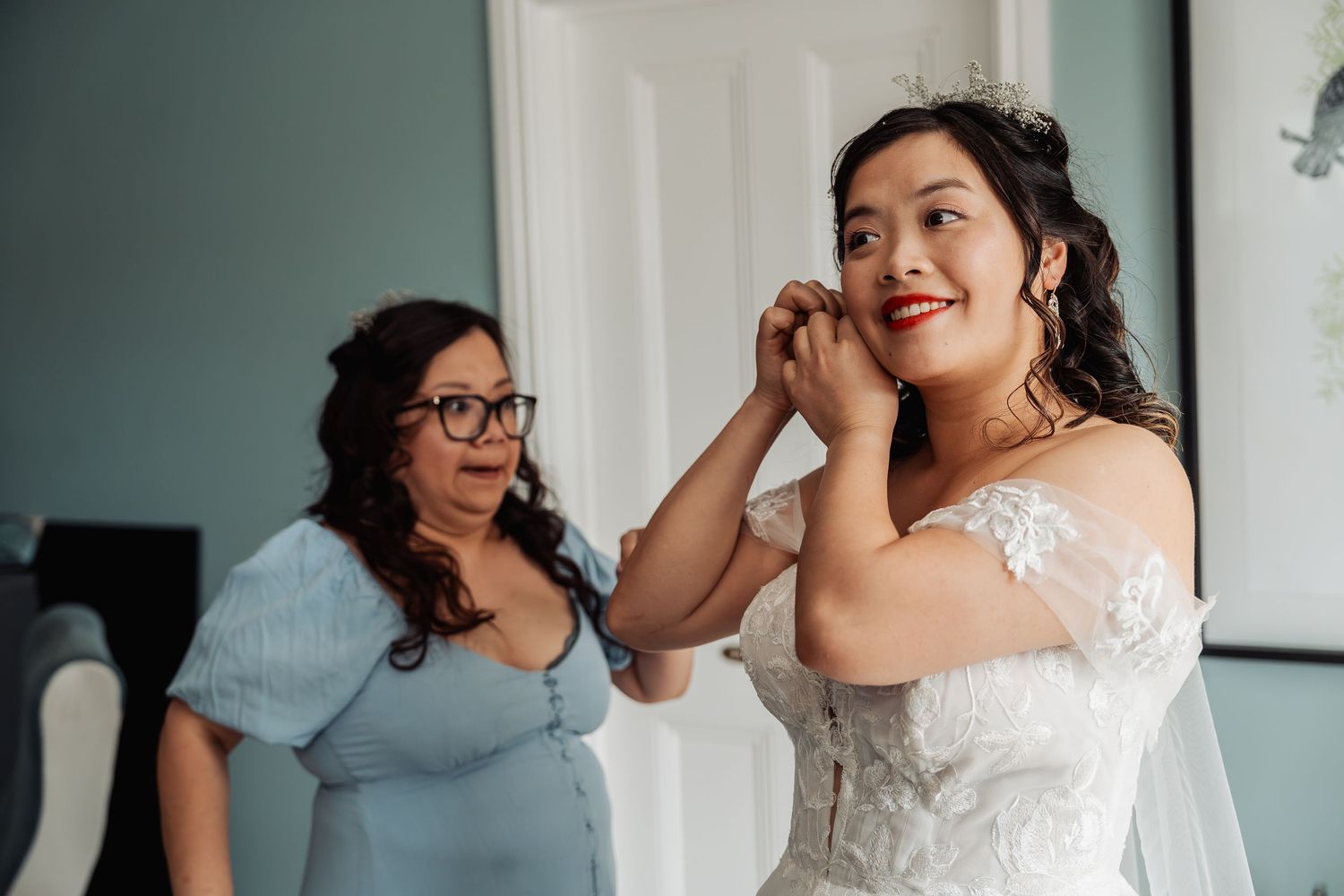 Bride in white lace gown puts on earrings while bridesmaid in blue dress watches during wedding preparations.