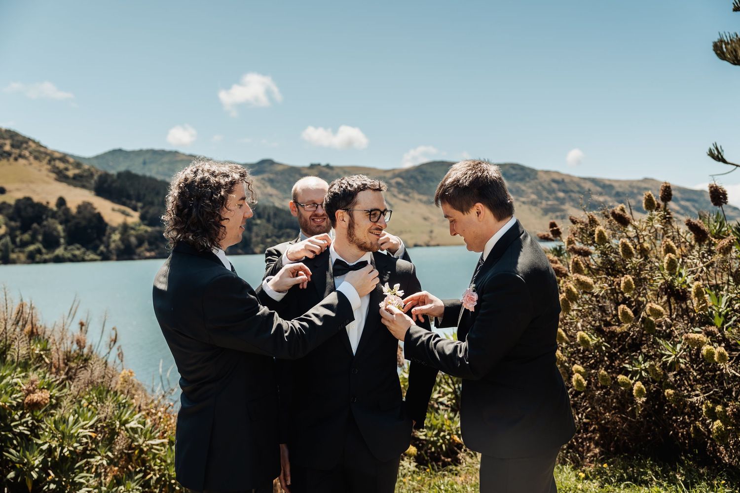 Groomsmen helping groom adjust bow tie outdoors near scenic lake and green hills on wedding day.