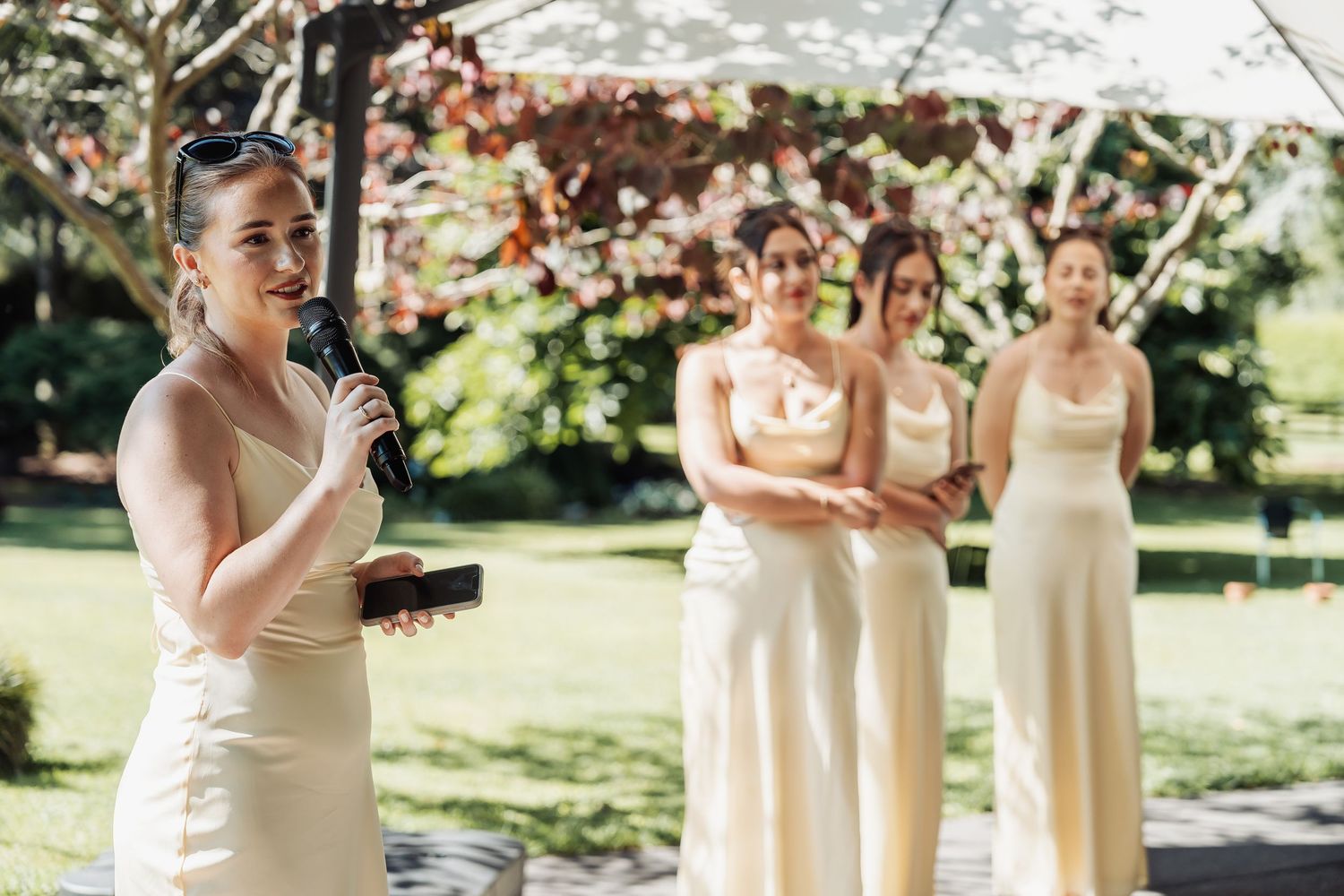 Bridesmaid in cream dress speaks into microphone at outdoor wedding ceremony while three bridesmaids stand in background.