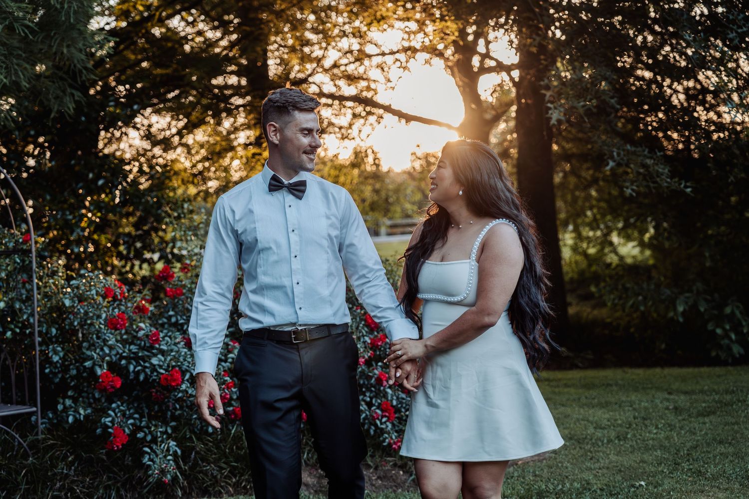 Couple holding hands walking in a garden at golden hour, man in bow tie and woman in white dress.