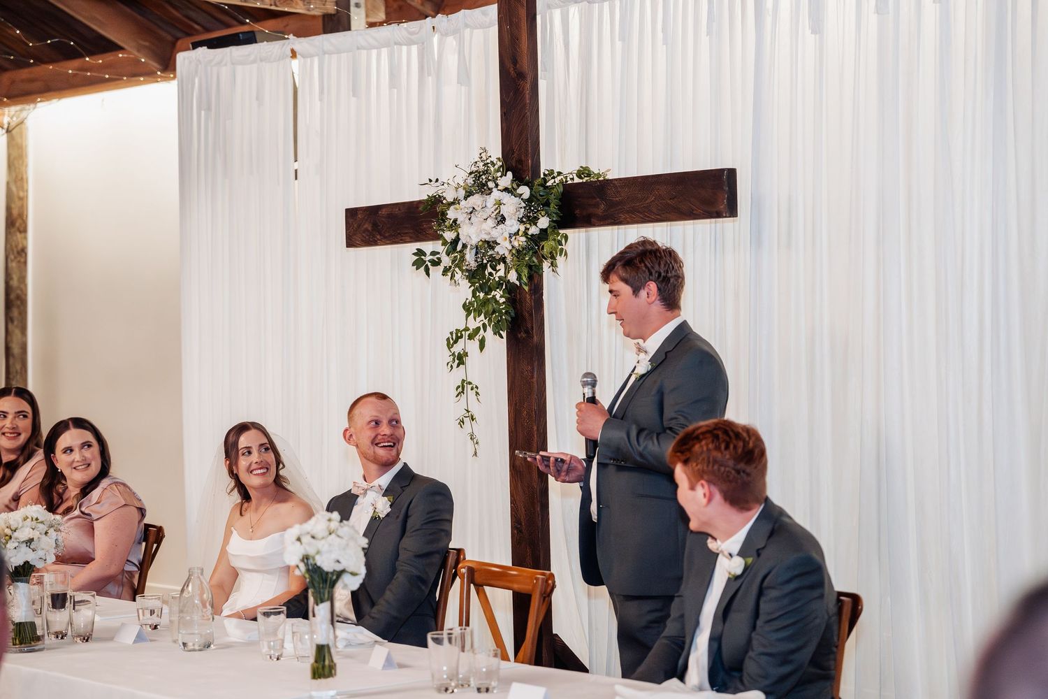 Best man delivering a heartfelt speech at wedding reception as bride and groom laugh at the head table.