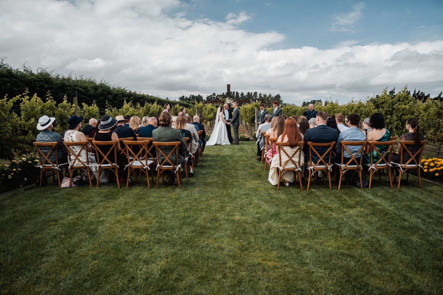 Outdoor wedding ceremony with guests seated on wooden chairs facing couple at vineyard altar.