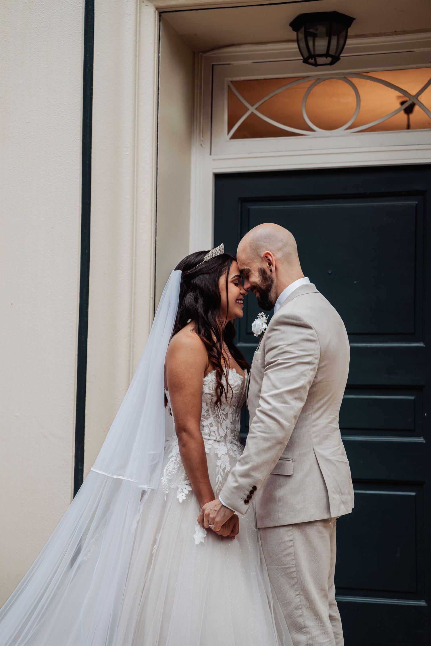 Bride and groom holding hands and touching foreheads romantically outside dark door after wedding.