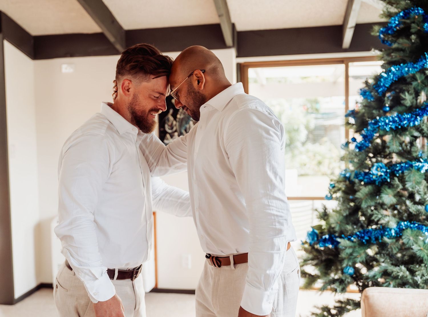 Two grooms share an intimate forehead touch moment in white shirts near a decorated Christmas tree.