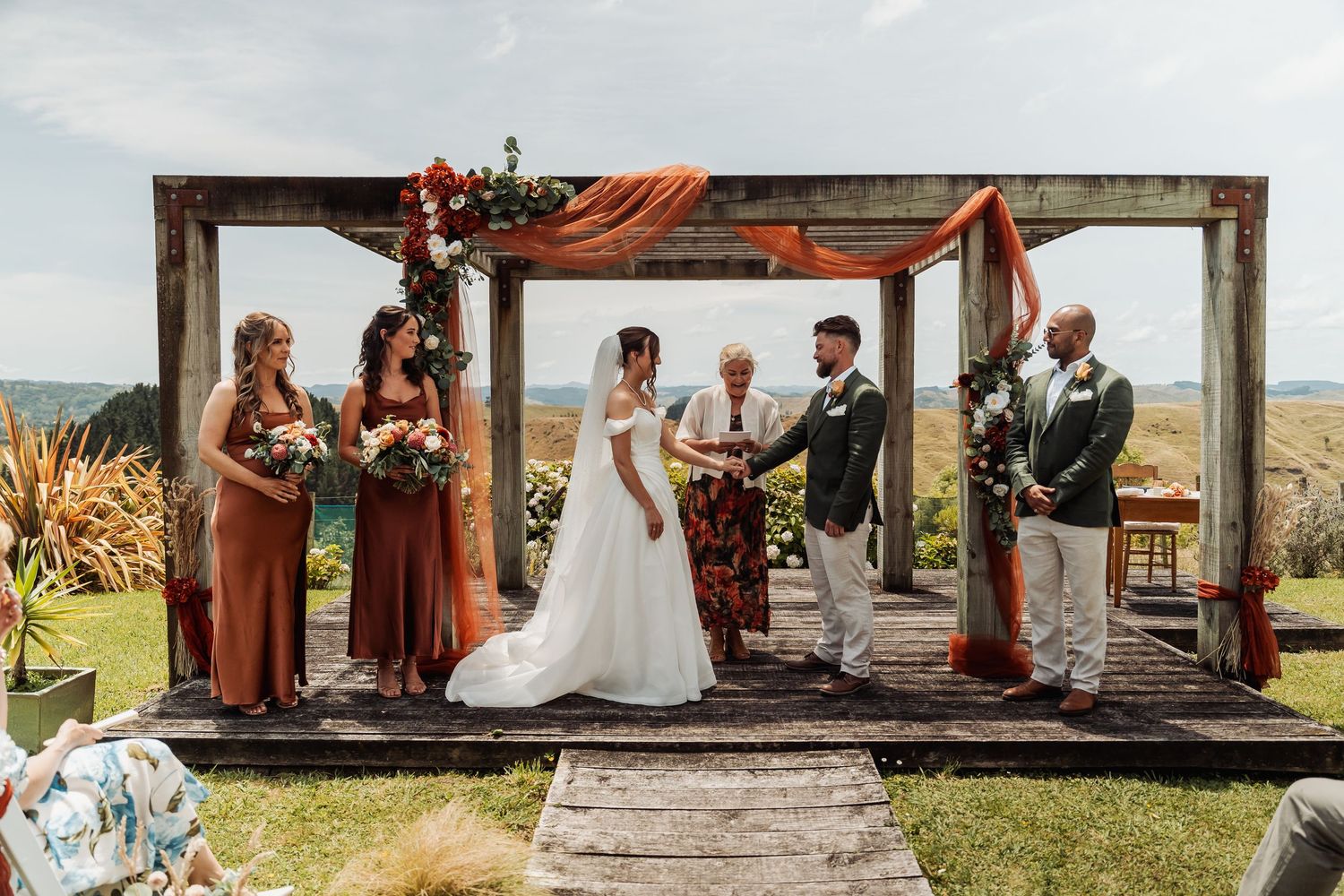 Bride and groom exchange vows under a rustic wooden arch with orange floral decor.