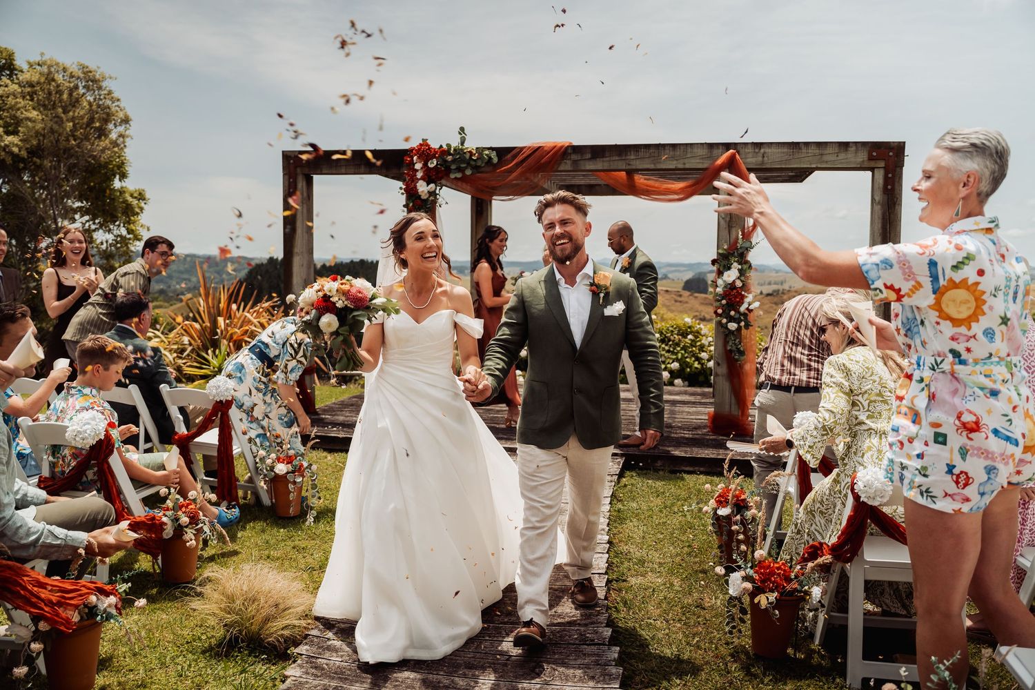 Newlyweds walking down aisle as guests throw confetti at outdoor wedding ceremony.