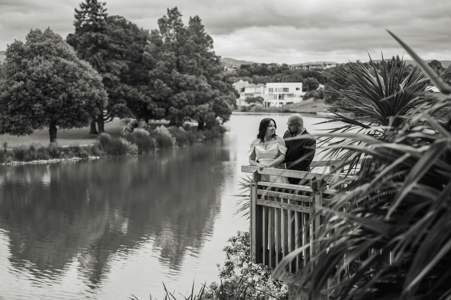 Black and white photo of bride and groom sitting together on a bridge over a lake.