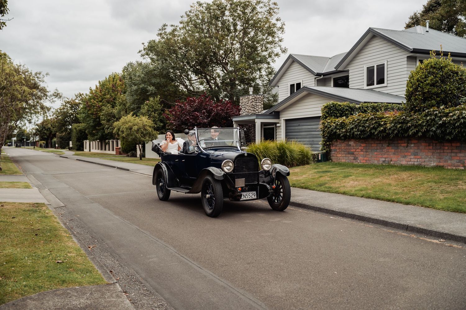Bride and groom riding in a vintage black classic car through a quiet suburban street on their wedding day.