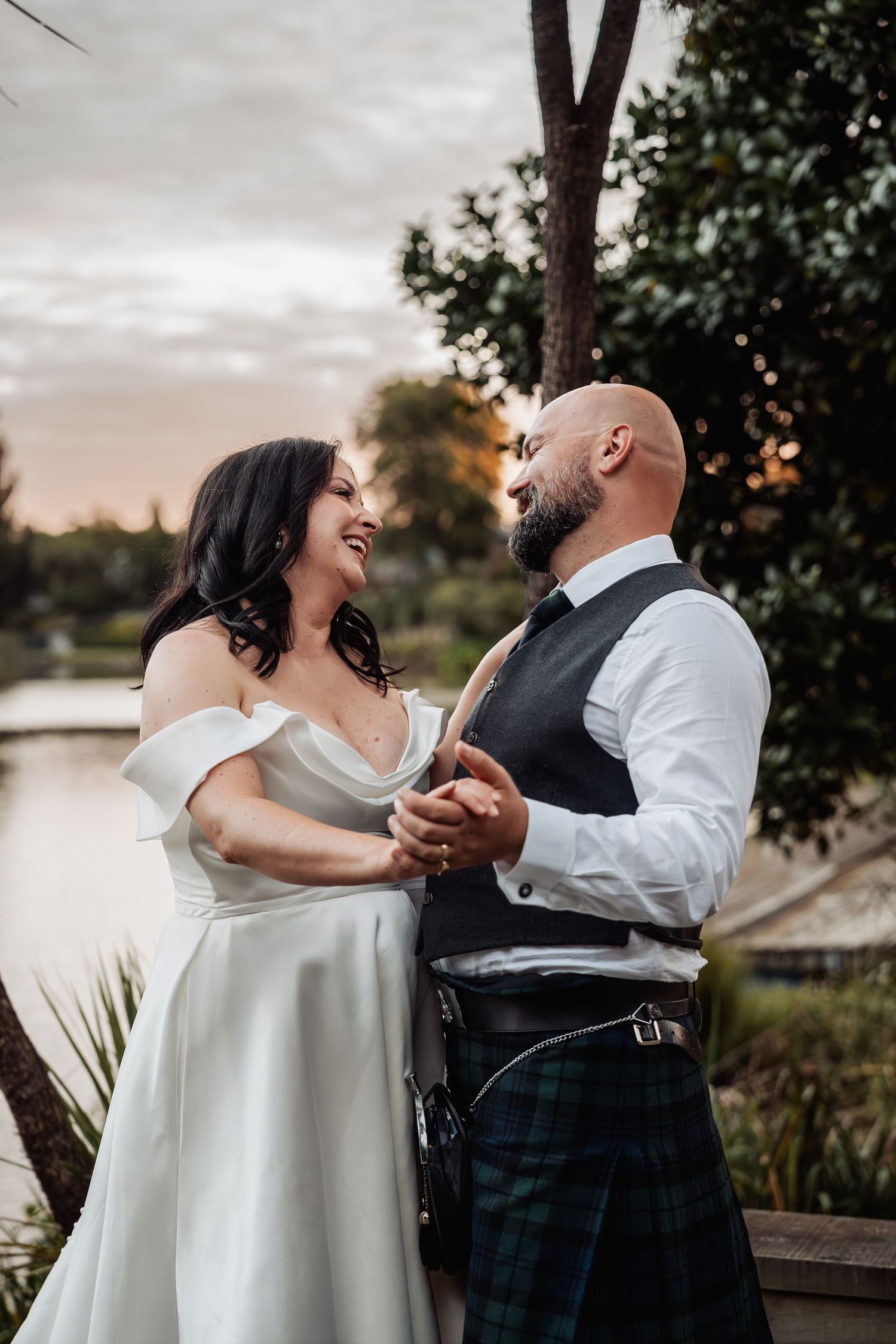 Bride and groom in kilt share a joyful moment holding hands near a tranquil lake at sunset.