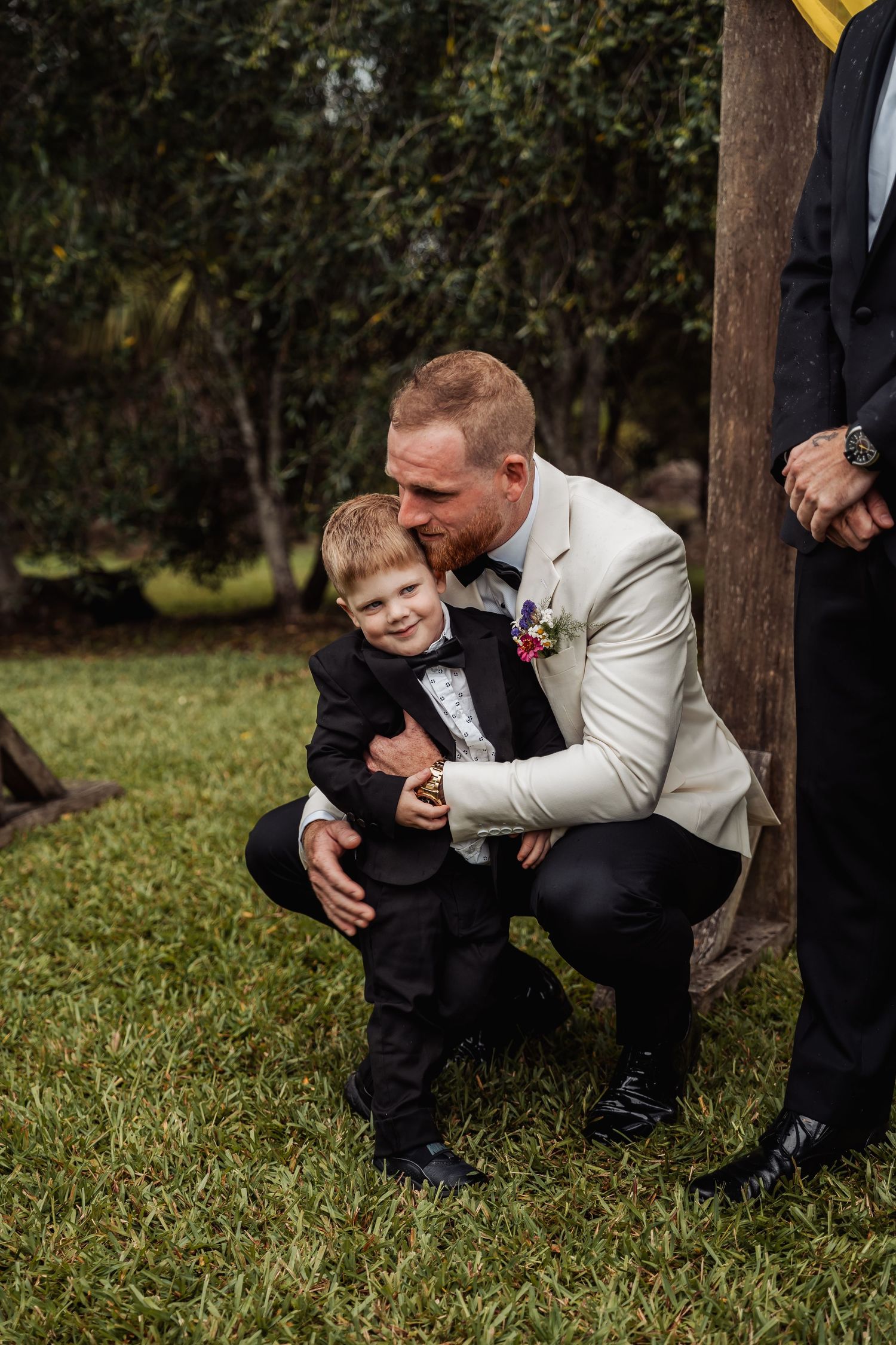 Groom in cream blazer crouching down to embrace a young boy in a black tuxedo.