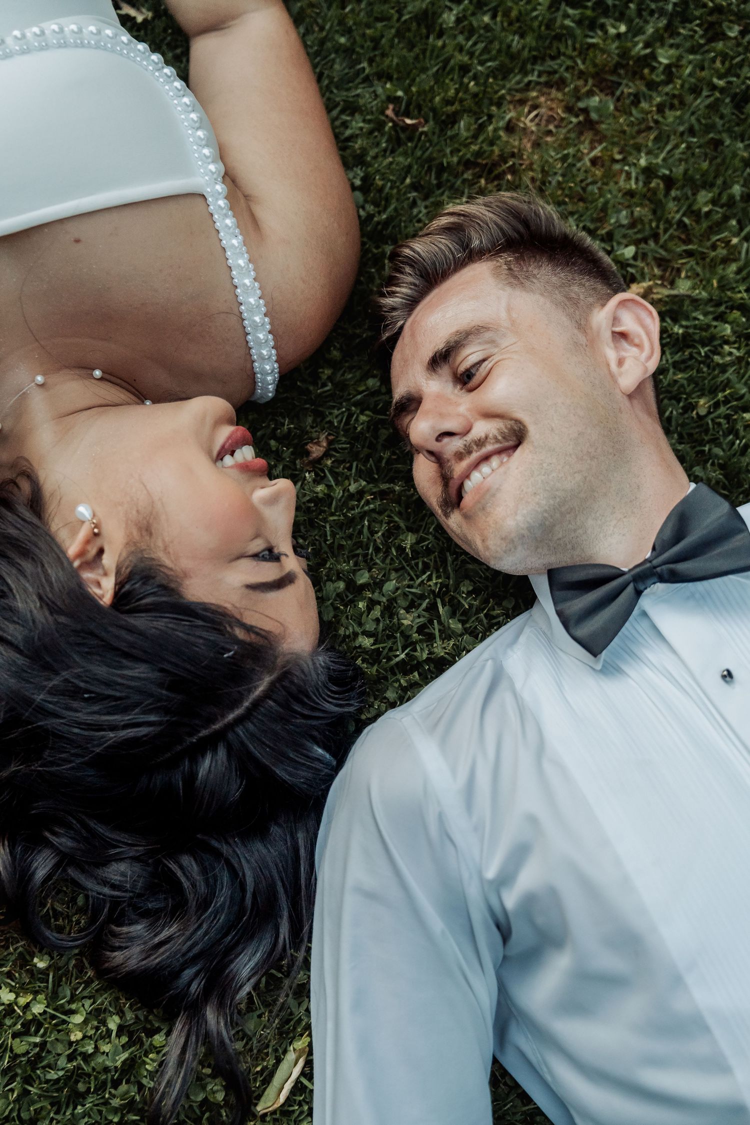 A smiling bride and groom in wedding attire lying on green grass, gazing at each other in a romantic outdoor setting.