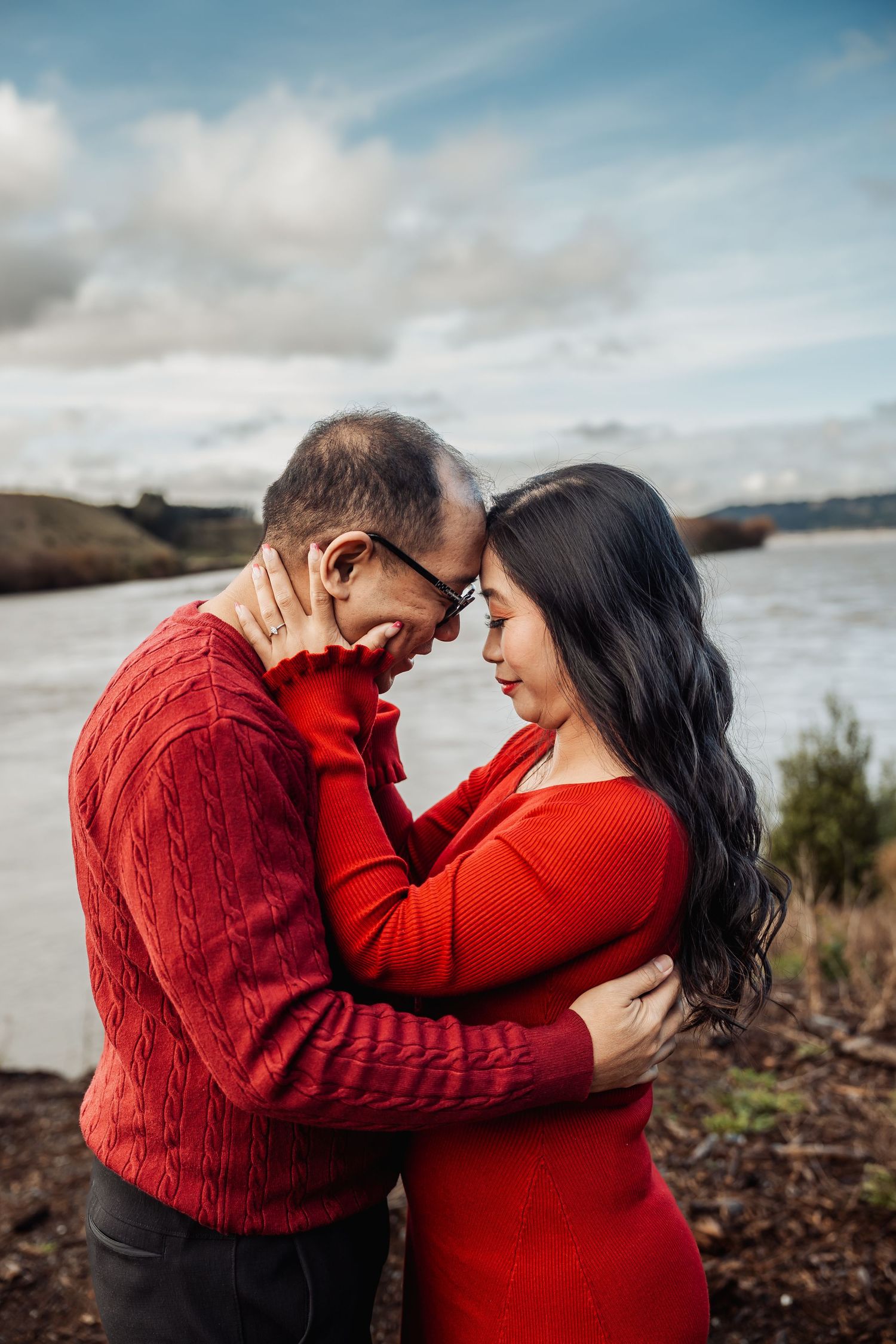 Couple in matching red outfits embrace forehead-to-forehead near a scenic waterway under a cloudy sky.