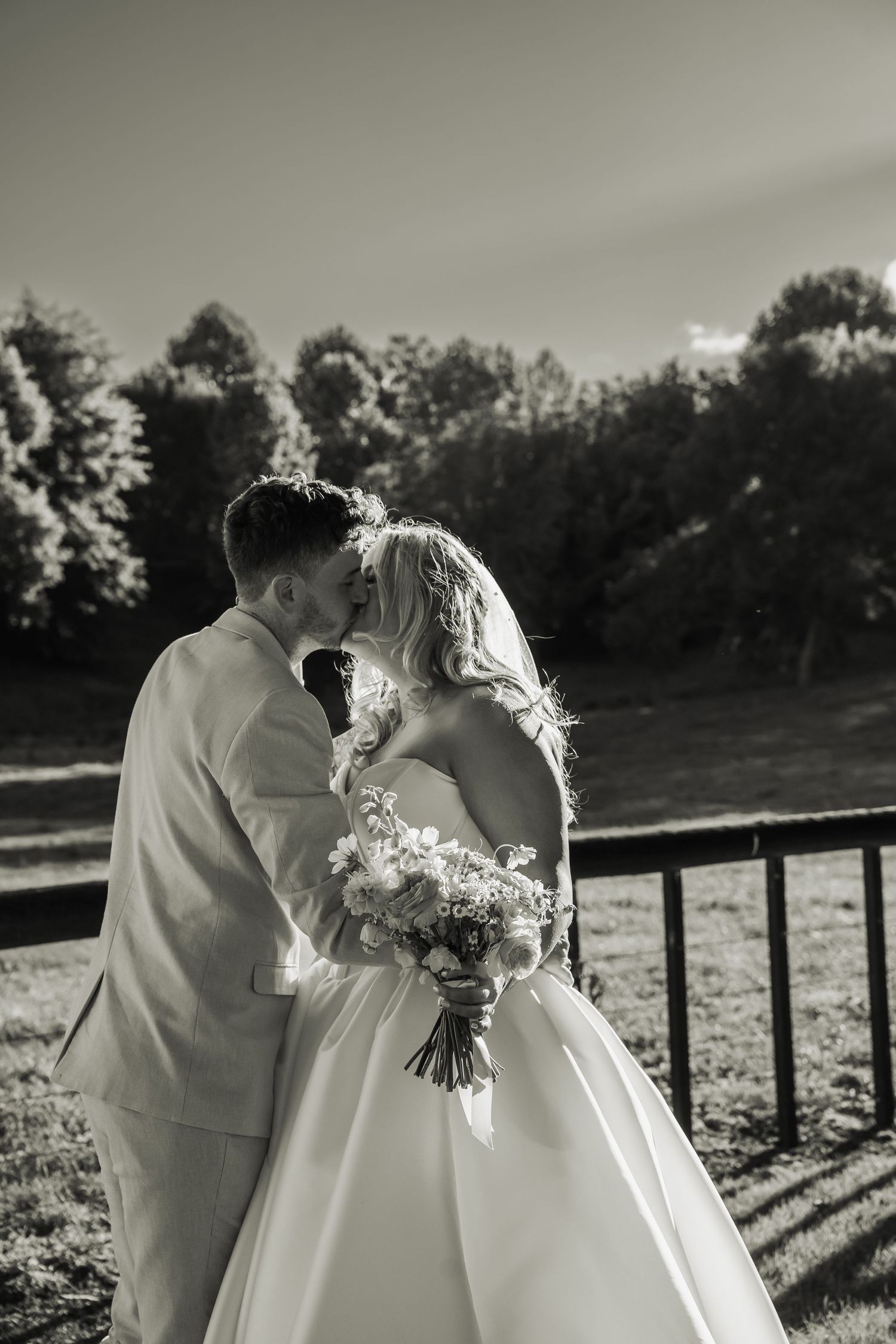 Black and white photo of bride and groom sharing a kiss outdoors with trees in background.