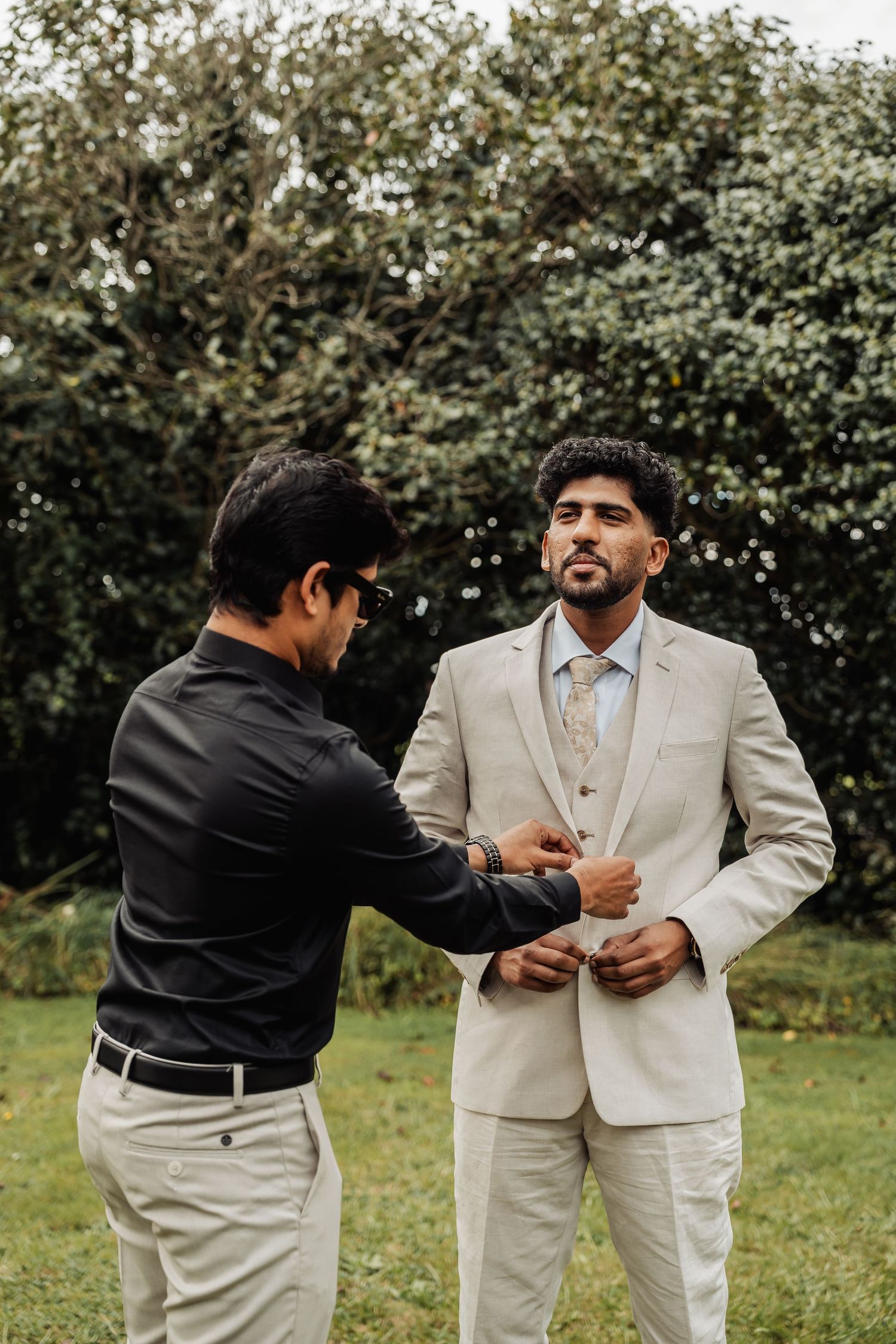 Groom in beige suit being helped by groomsman outdoors with lush green trees in the background.