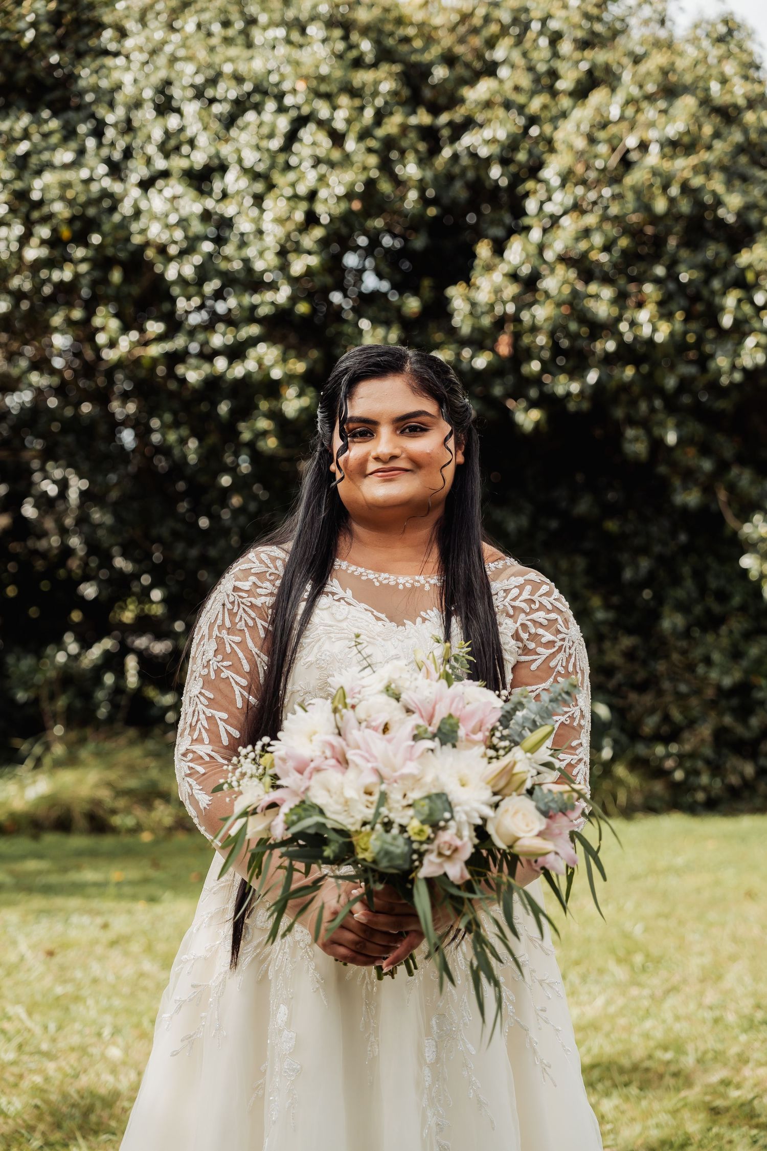 Bride in lace wedding dress holds pink and white floral bouquet smiling outdoors on wedding day.