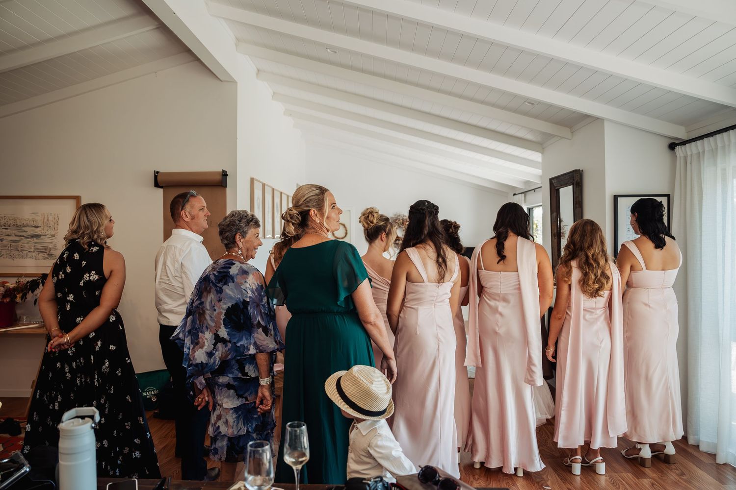 Bridesmaids in pink dresses and guests gathered indoors watching a wedding preparation moment.