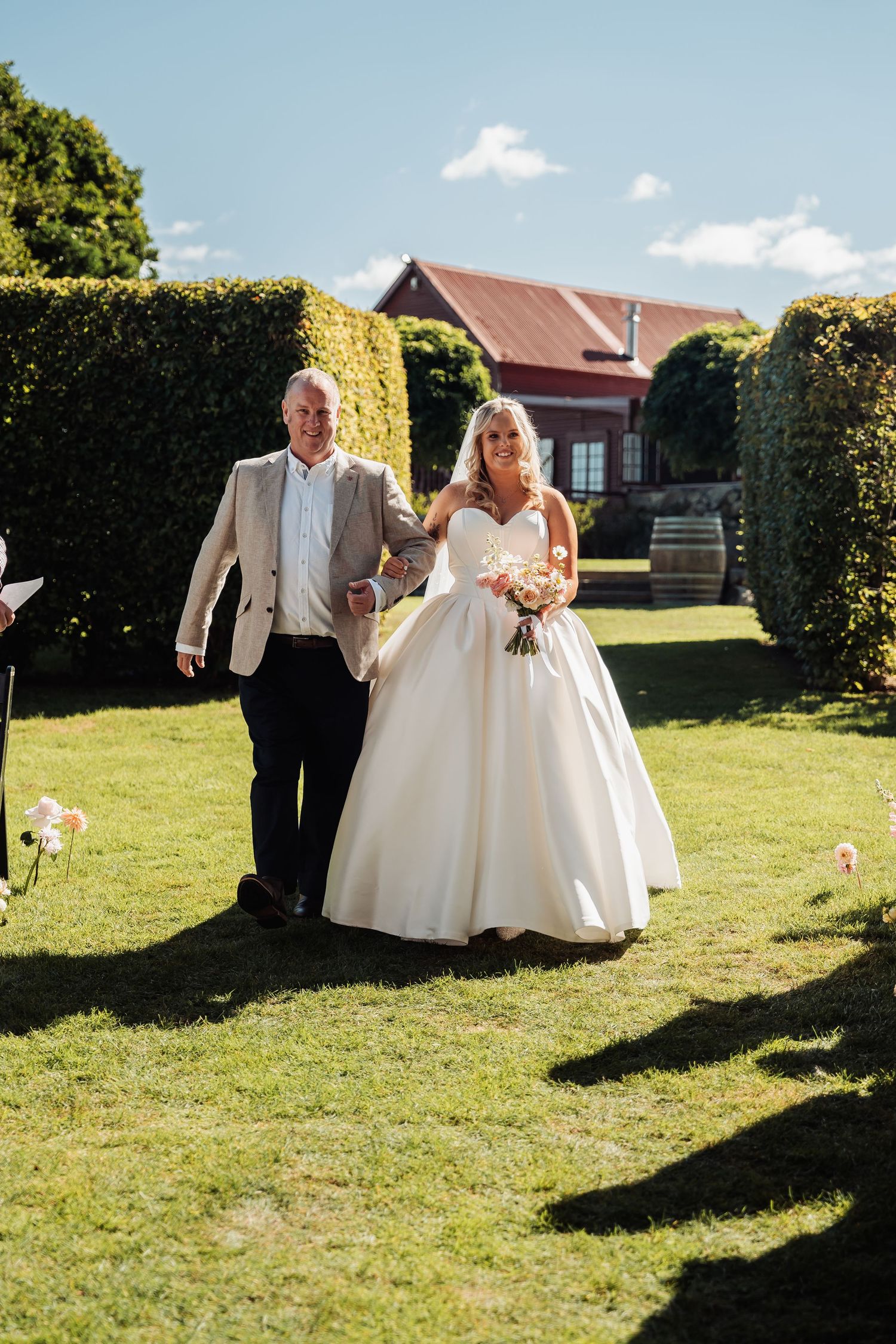 Bride in white ballgown walks arm-in-arm with escort through garden ceremony aisle.