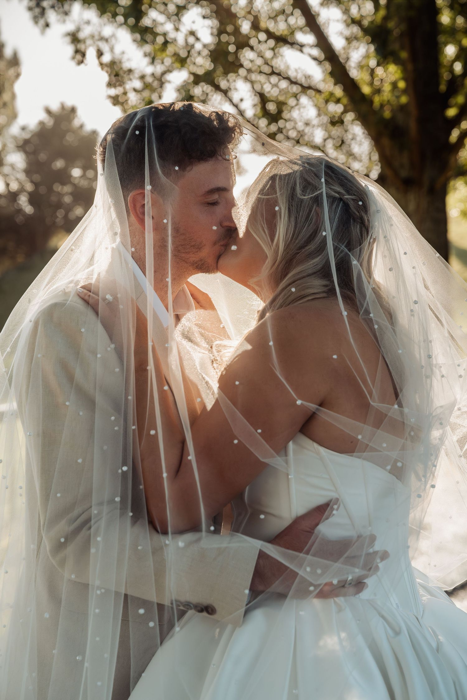 Bride and groom share a romantic kiss under a pearl-embellished wedding veil in soft sunlight.