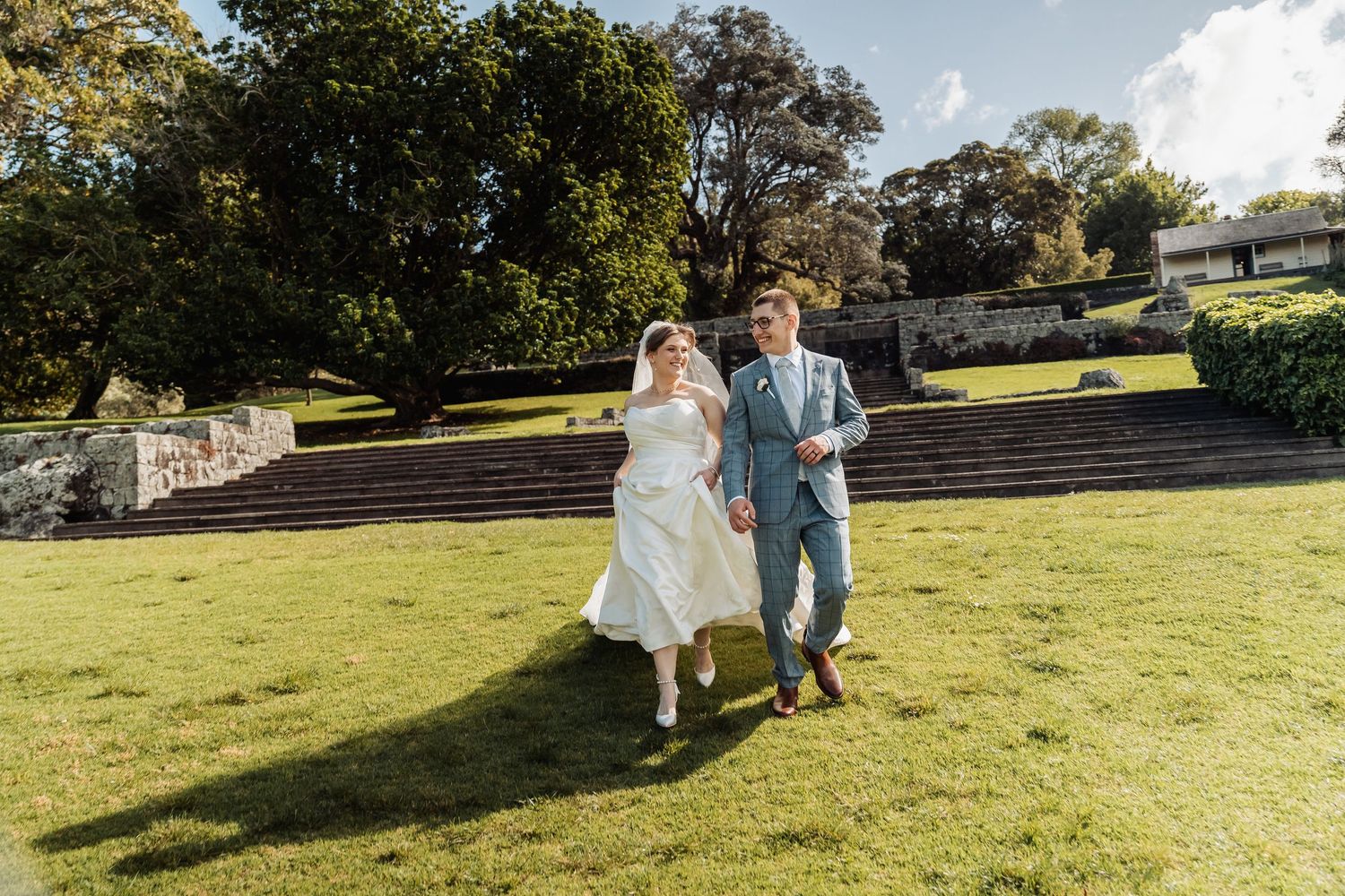 A bride and groom hold hands while walking across a lush green lawn at an outdoor wedding venue.