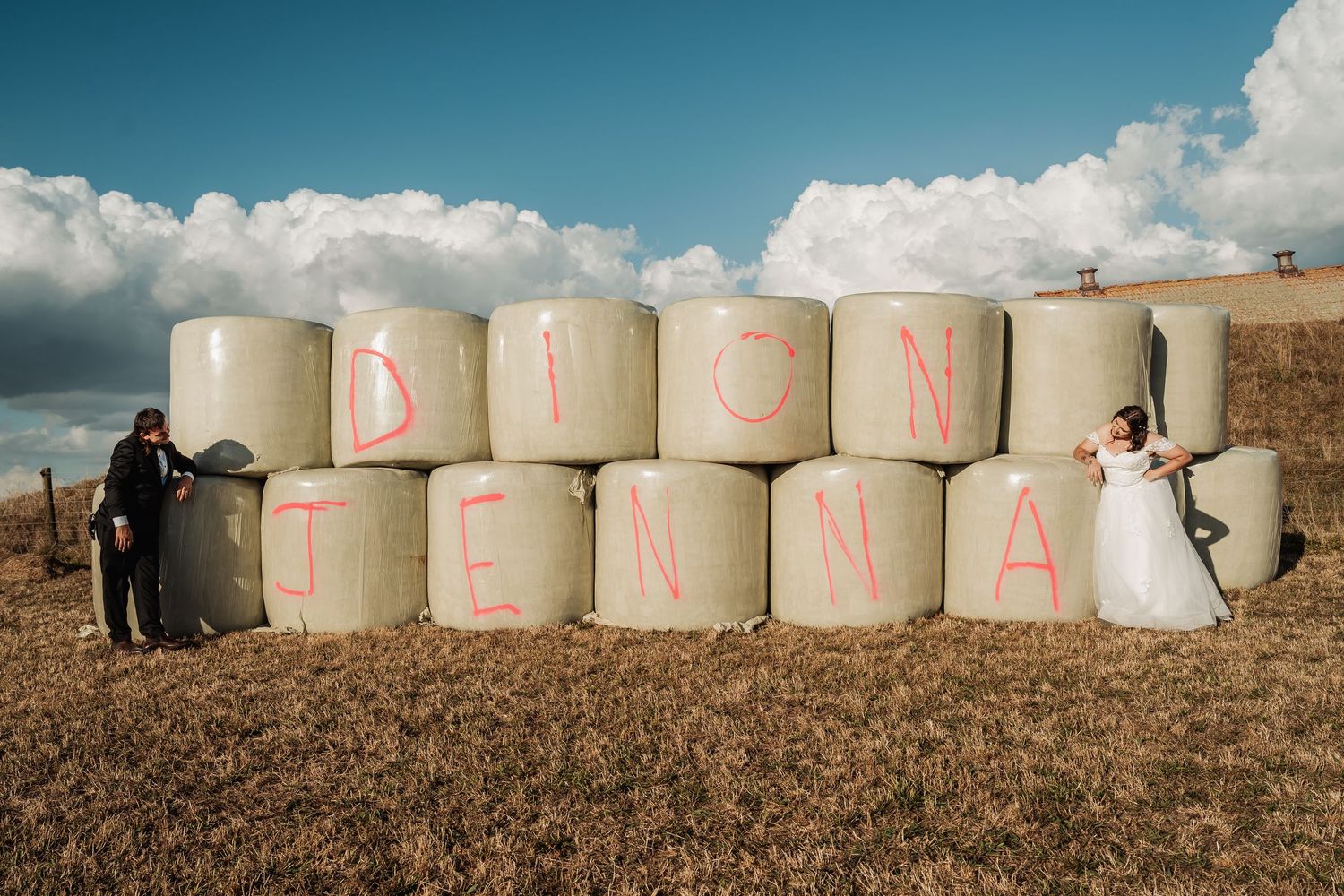 Bride and groom posing beside hay bales spelling out DION and JENNA at a farm wedding.