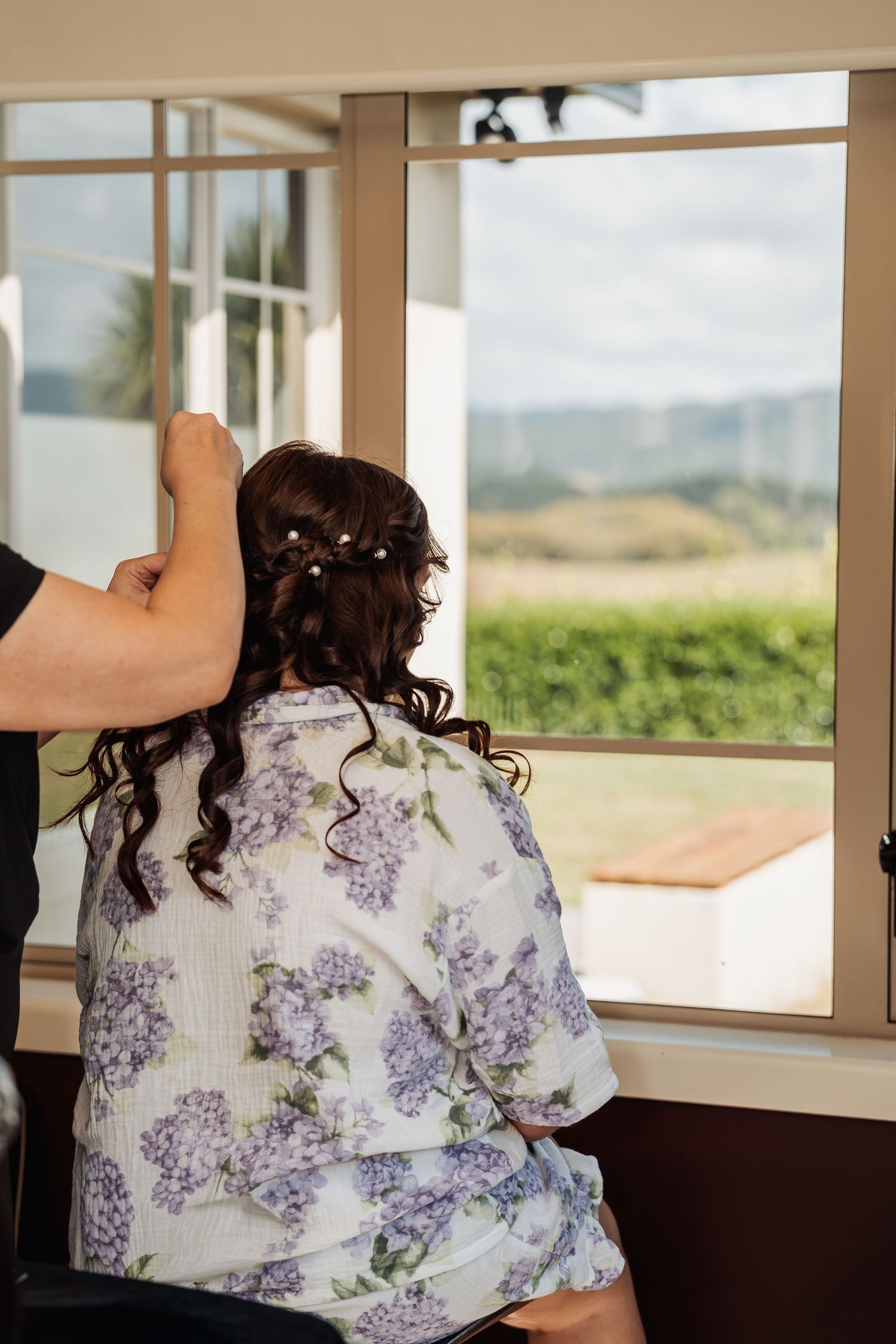 Hairstylist adding pearl pins to brunette bride's elegant updo while she sits in floral robe near window.