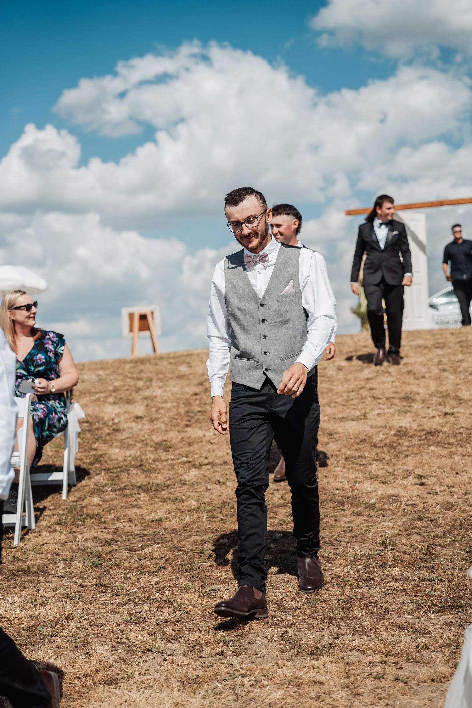 Smiling groom in grey vest and bow tie walks down outdoor wedding aisle under blue cloudy sky.