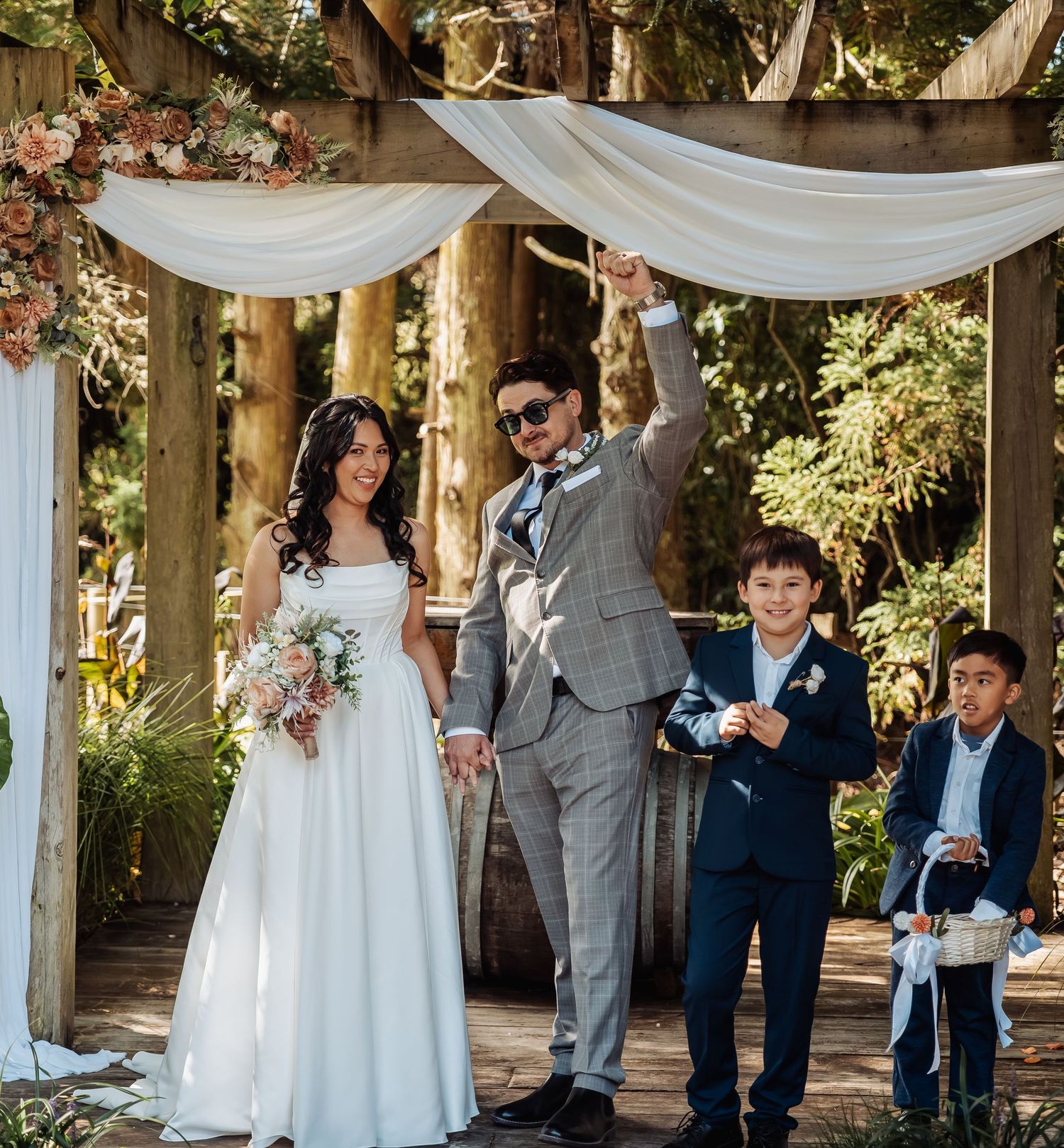 Groom raises fist in celebration beside bride in white gown and two boys at outdoor wedding ceremony.