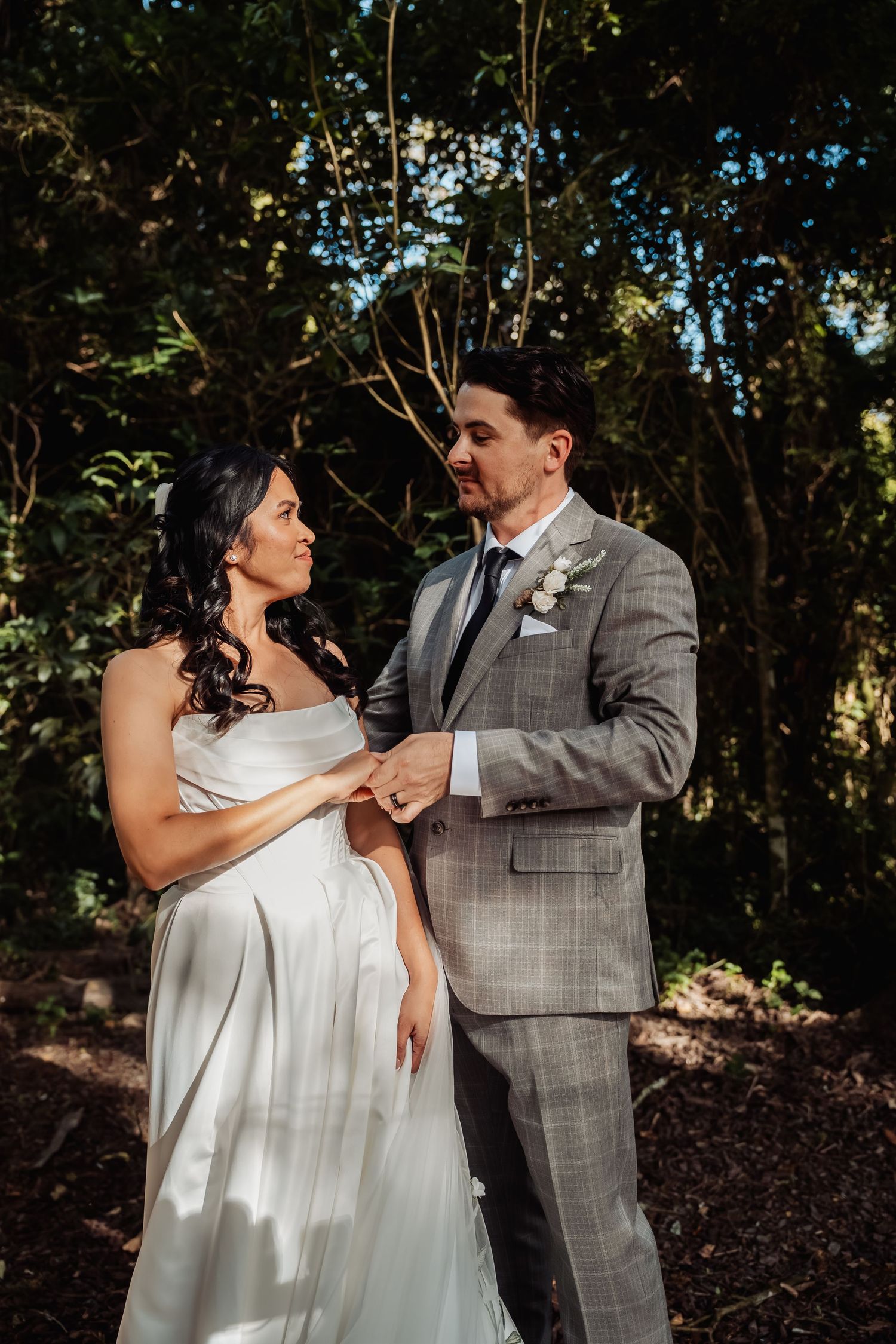 Bride in white gown and groom in plaid suit hold hands, gazing at each other in a sunlit forest setting.