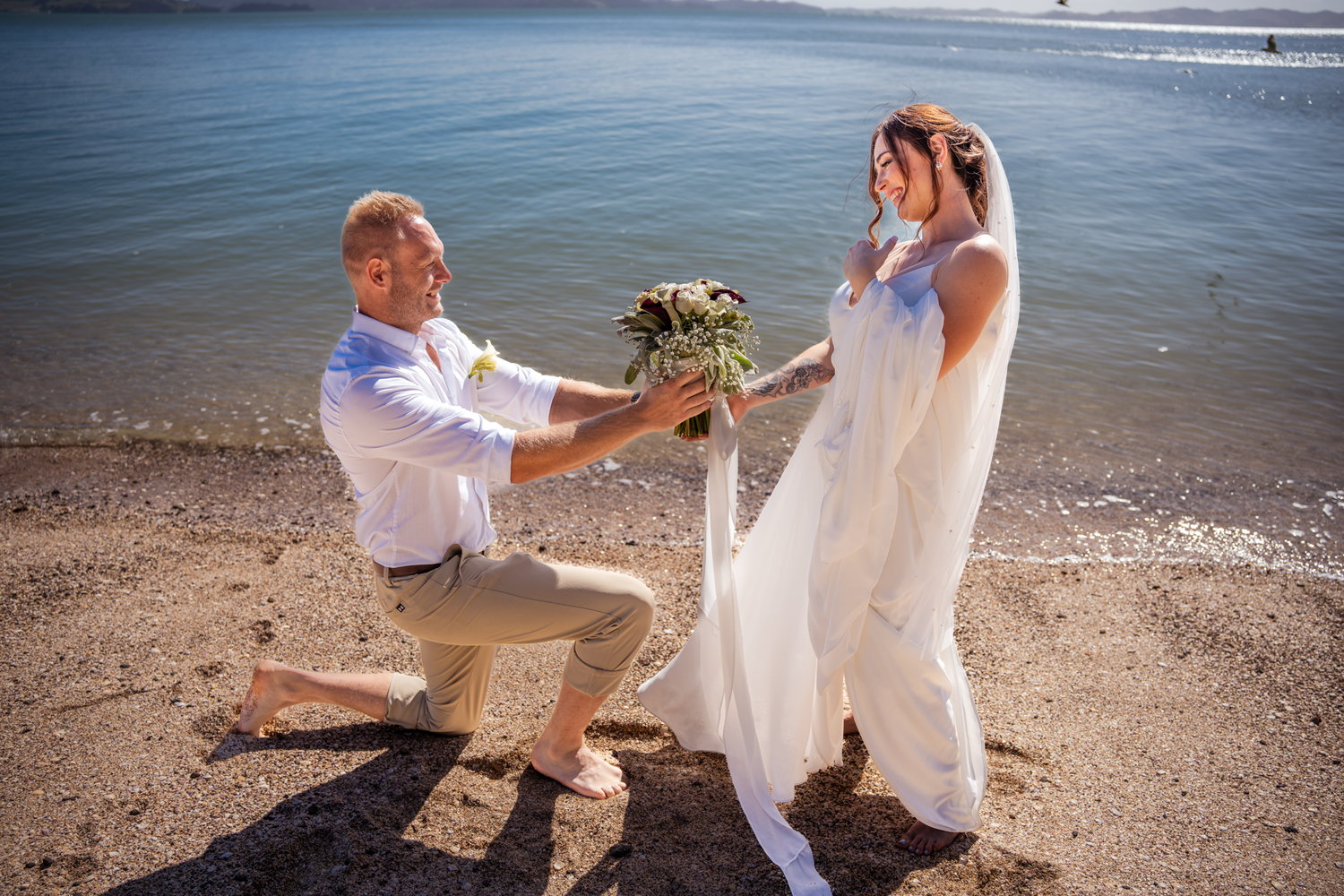 Romantic beach proposal scene with white dress and bouquet against ocean backdrop.