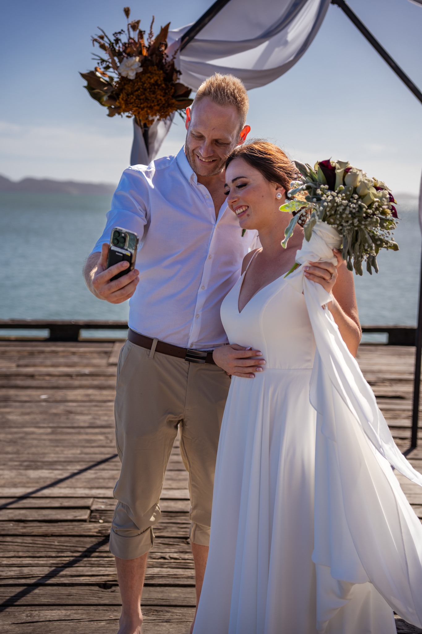A happy couple takes a selfie together on a wooden pier overlooking the ocean.