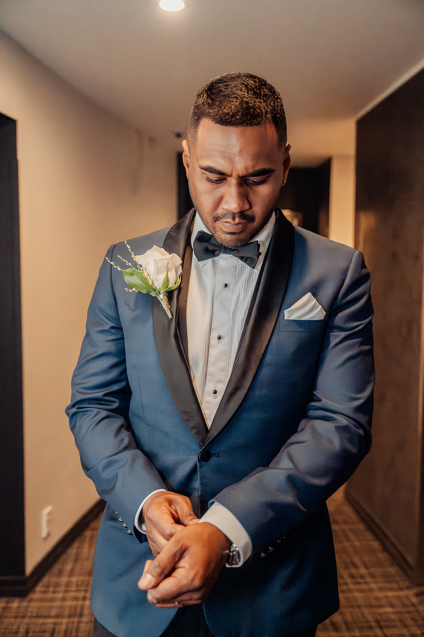 Groom in blue tuxedo adjusts cufflinks while standing in hotel hallway.