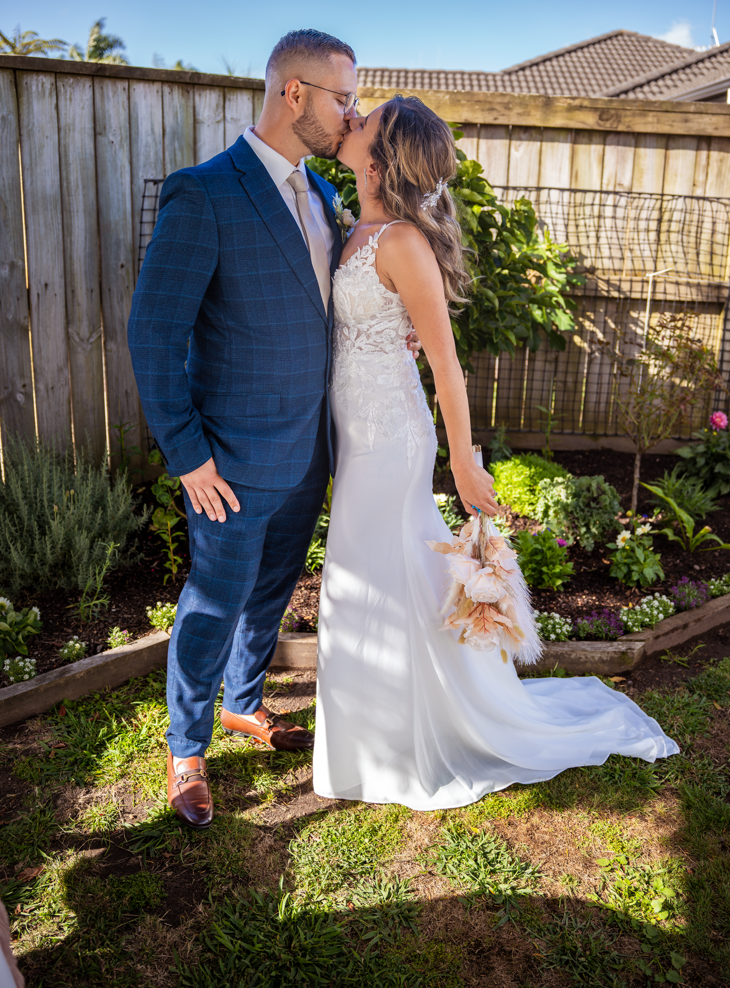 A romantic wedding couple poses in a backyard garden with a wooden fence and flowering plants.
