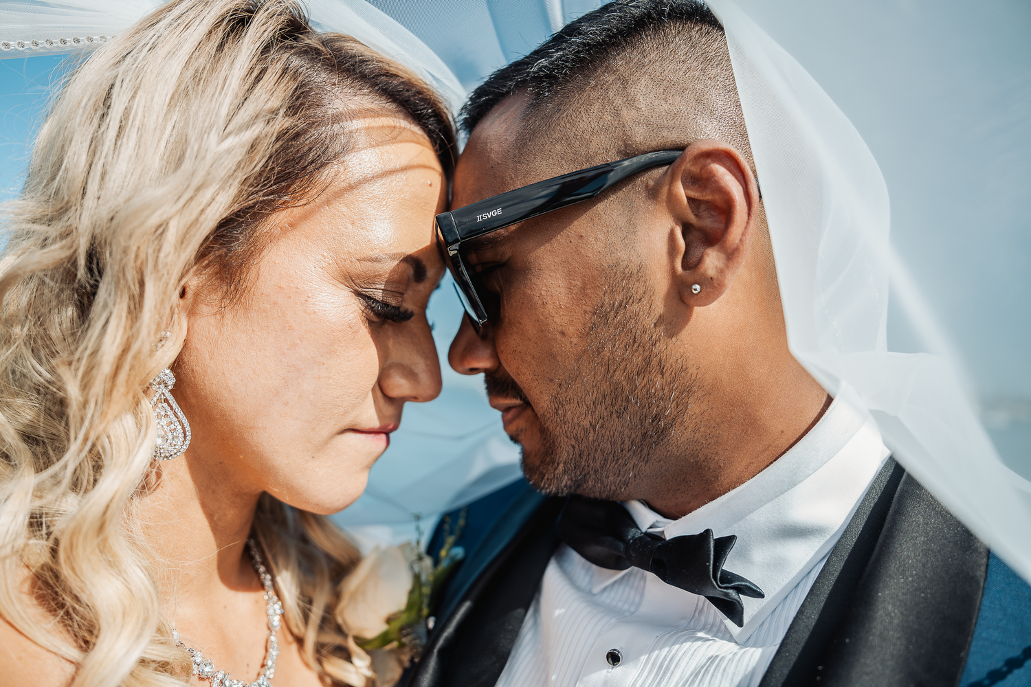 An intimate moment between newlyweds touching foreheads against a blue sky background.