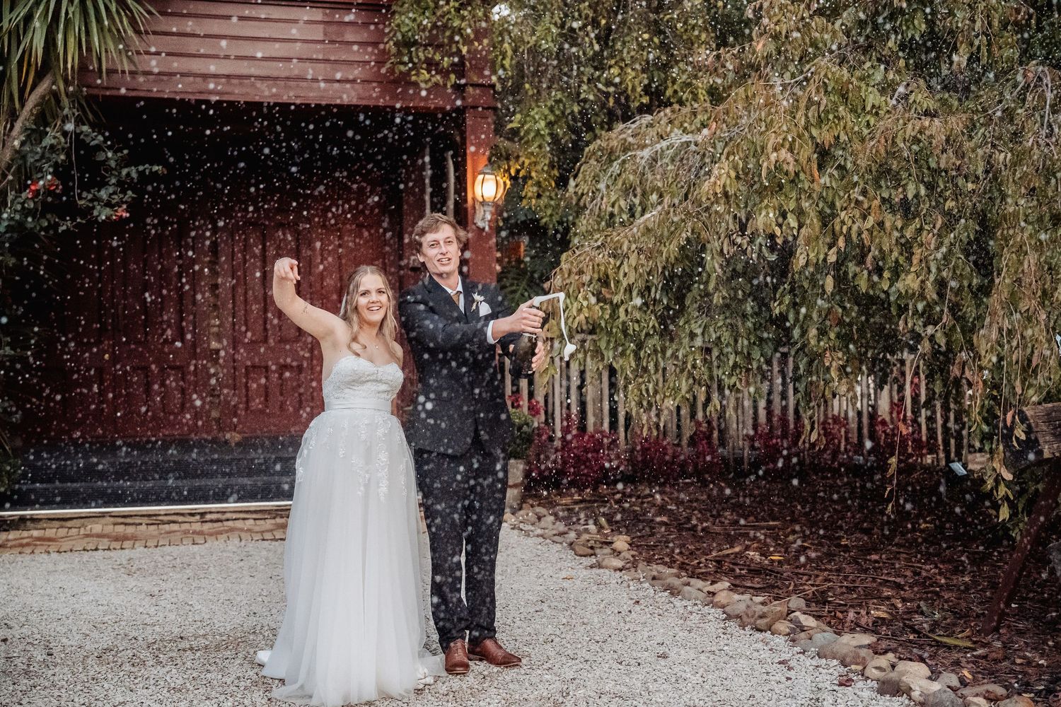 Couple celebrates joyfully on their wedding day with confetti falling around them outside a rustic red building.