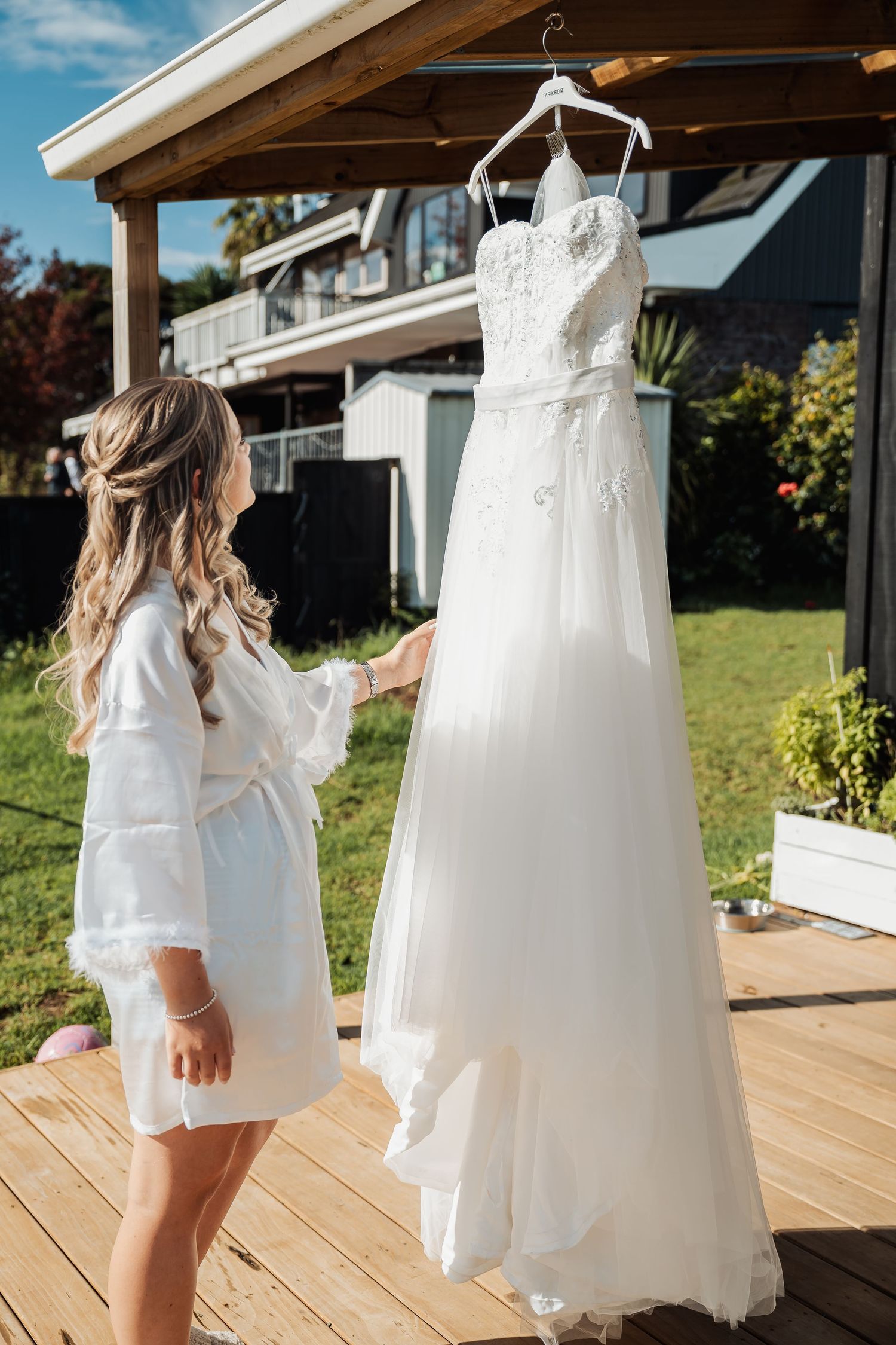 A white wedding dress hangs on a wooden porch as someone in a white robe reaches towards it on a sunny day.