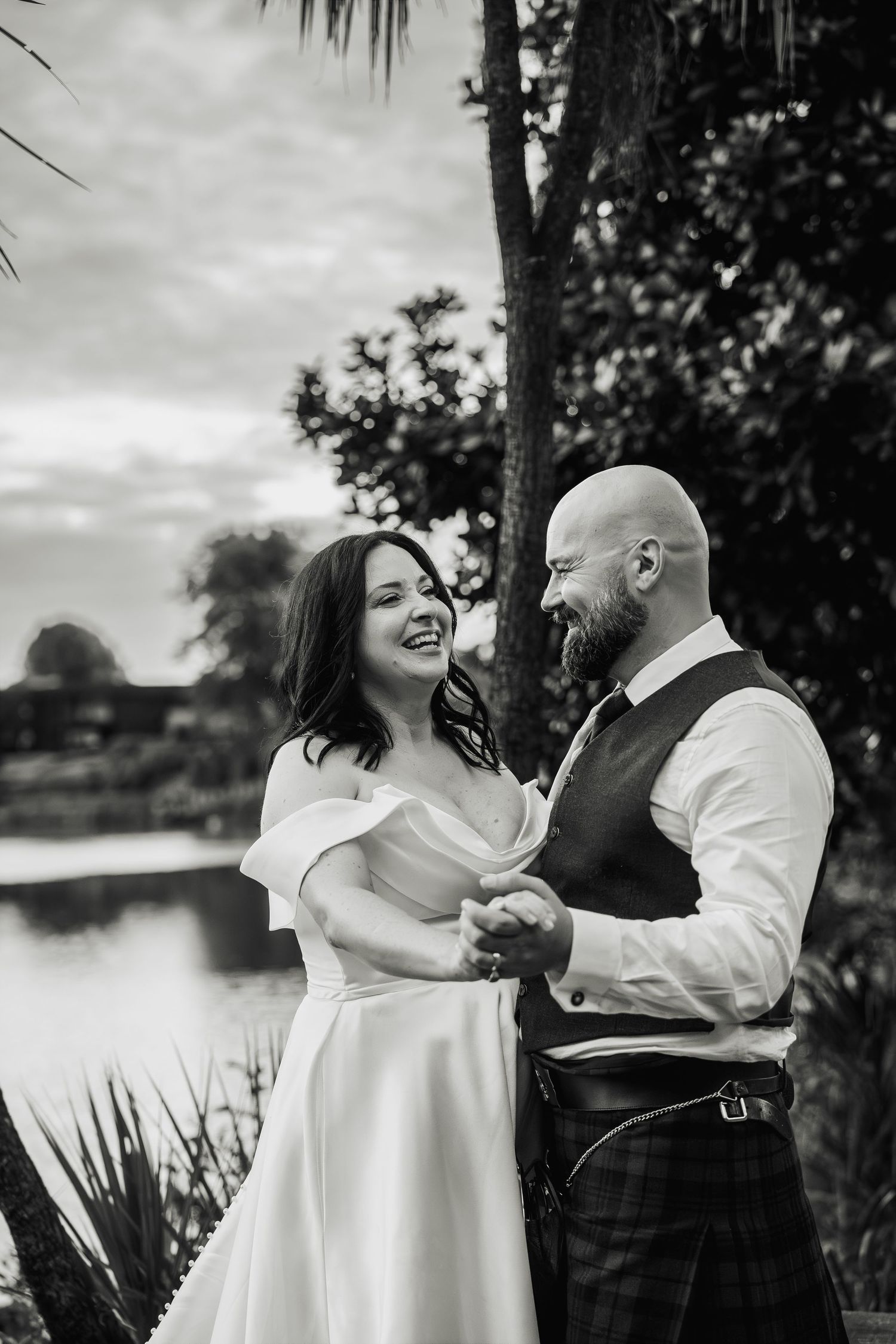 Black and white photo of laughing bride and bearded groom holding hands near lake with palm trees.