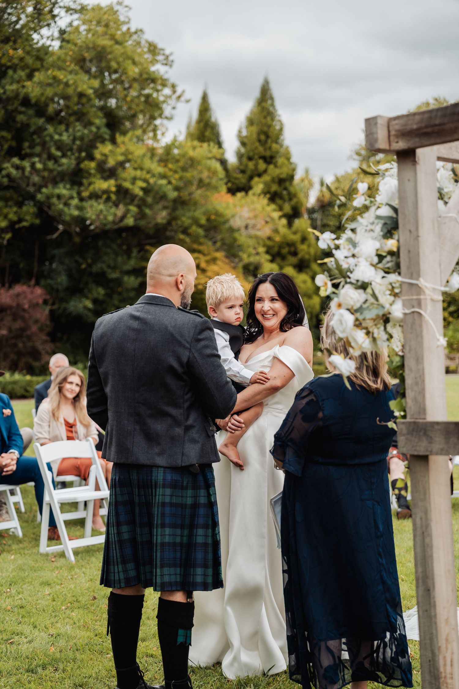 Bride and groom in tartan kilt hold a young child during an outdoor wedding ceremony with seated guests.