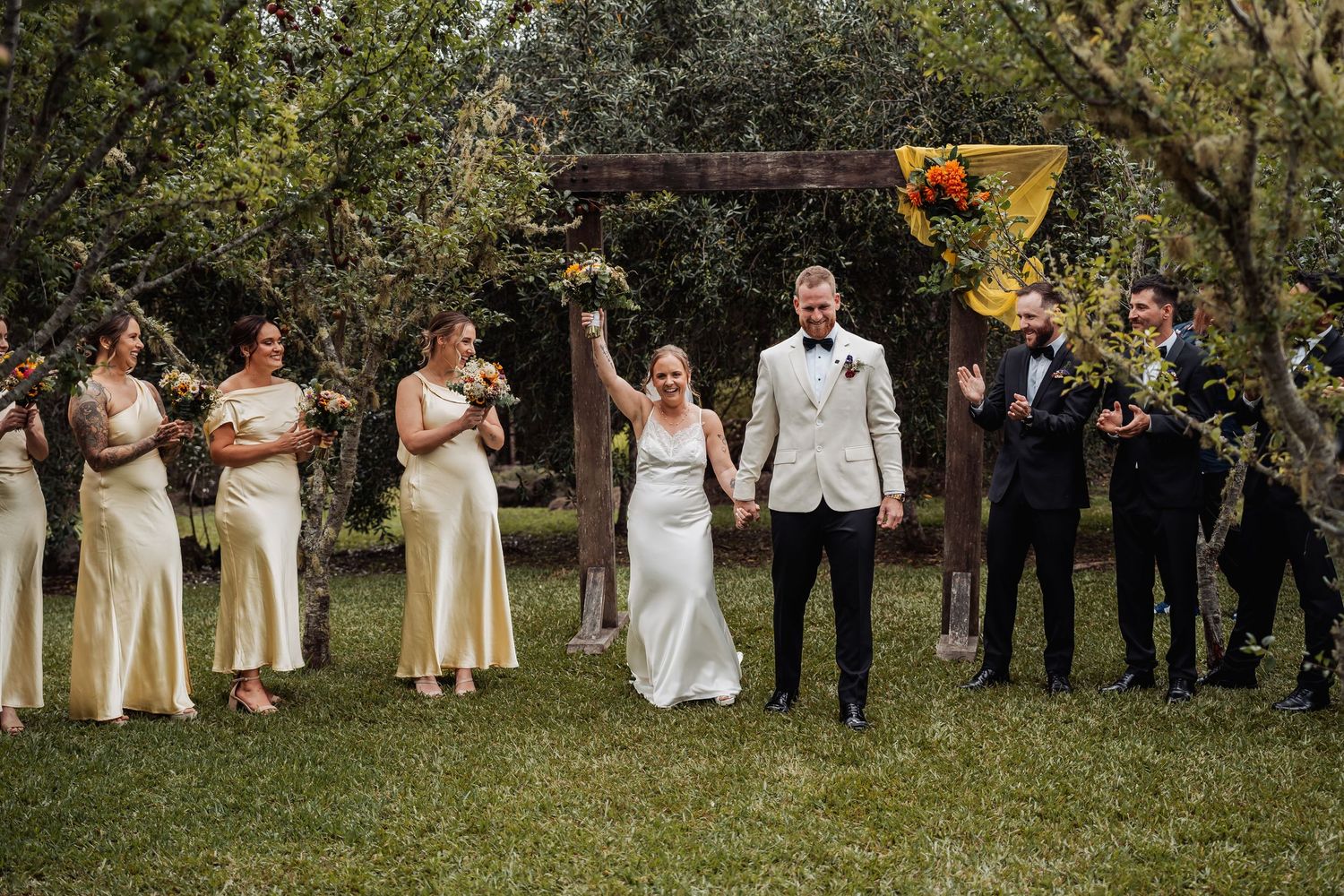 Wedding party celebrating outdoors with bridesmaids in yellow satin dresses and groomsmen under a floral arch.