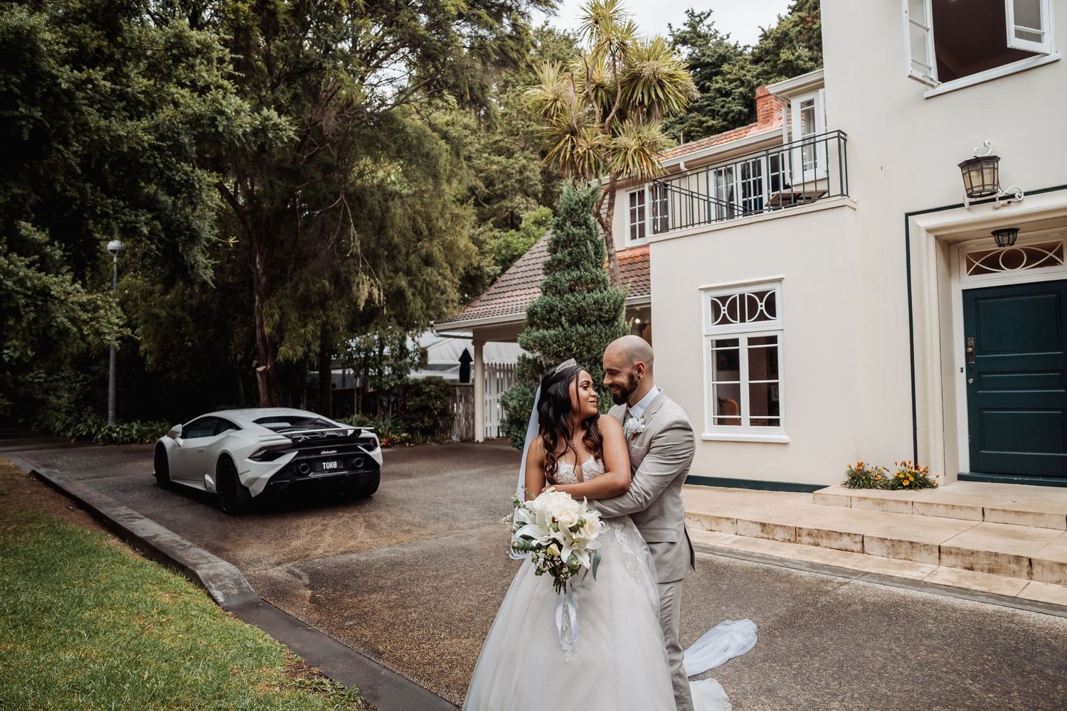Bride and groom embracing outside elegant venue with white Lamborghini in background.