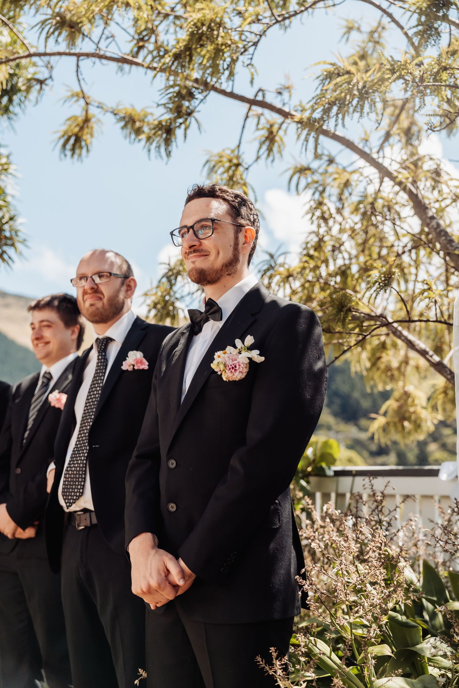 Groom and groomsmen in black suits with floral boutonnieres standing outdoors at wedding ceremony.