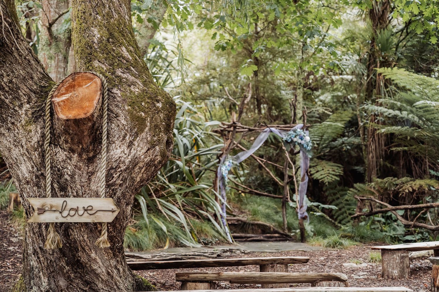 Rustic woodland wedding ceremony setup with a love sign on a tree and decorated arch among lush ferns.