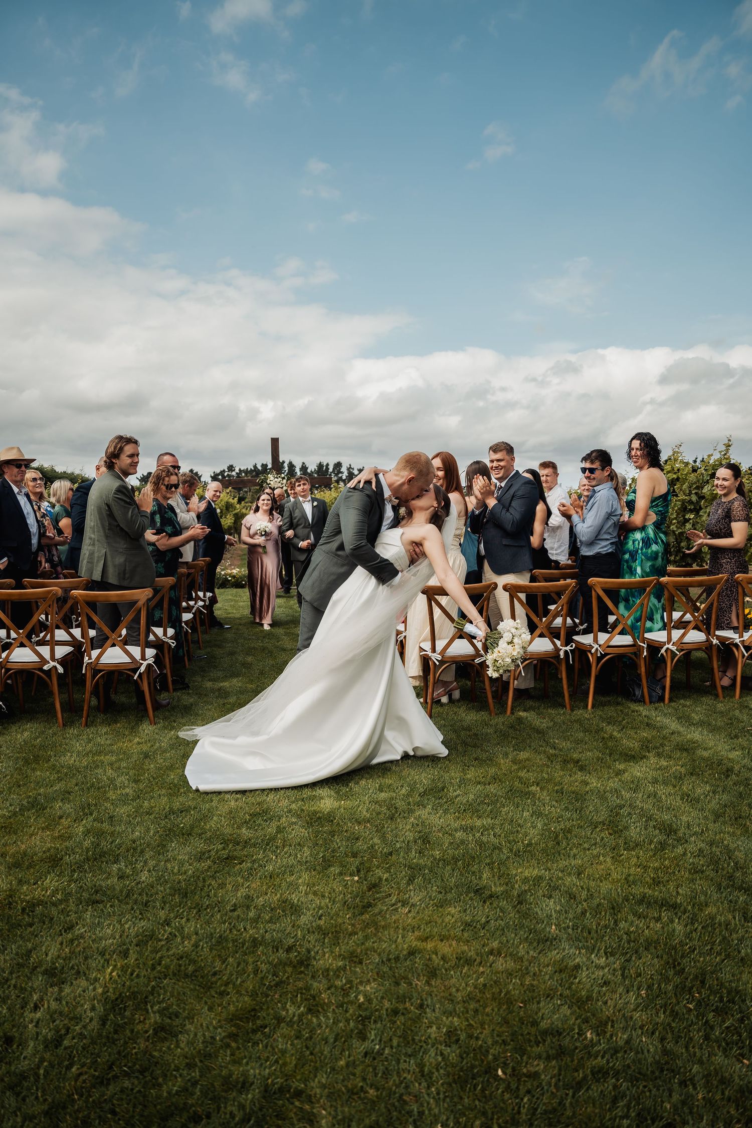 Groom dips bride for a romantic kiss while wedding guests cheer along the aisle at an outdoor ceremony.