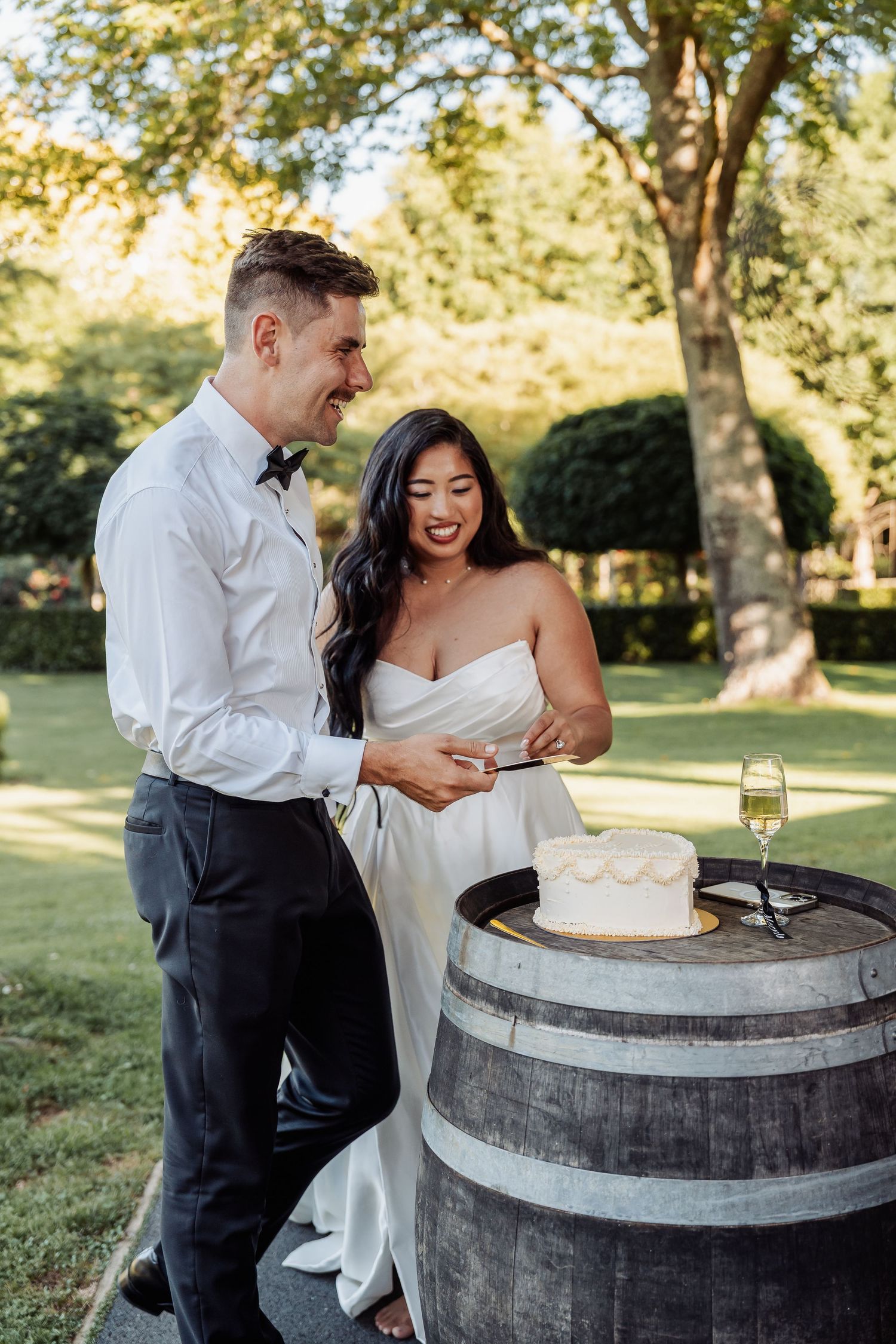 Smiling bride and groom cutting wedding cake on a rustic wine barrel outdoors.