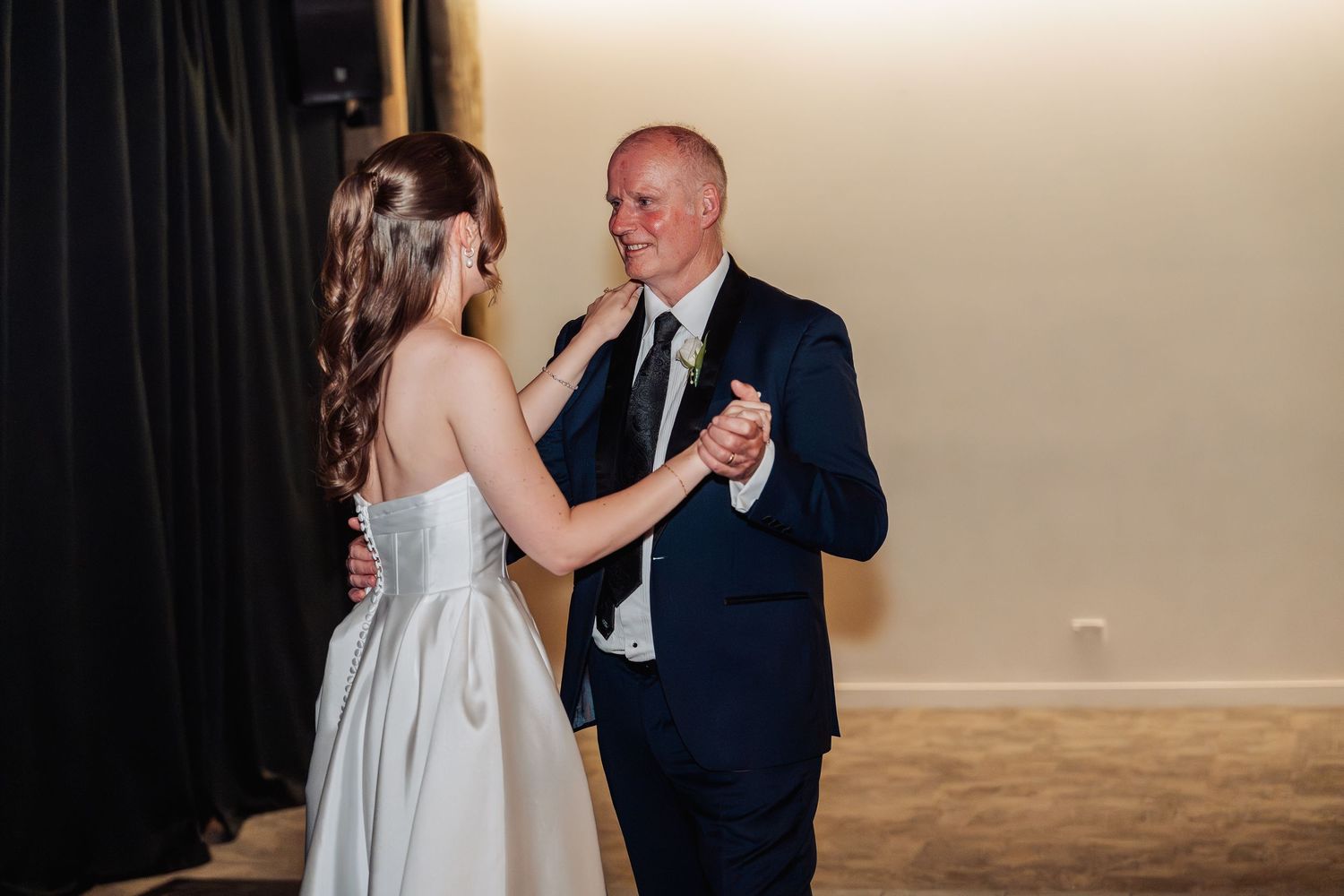 Bride and groom share an intimate first dance, smiling warmly at each other in a softly lit wedding reception venue.