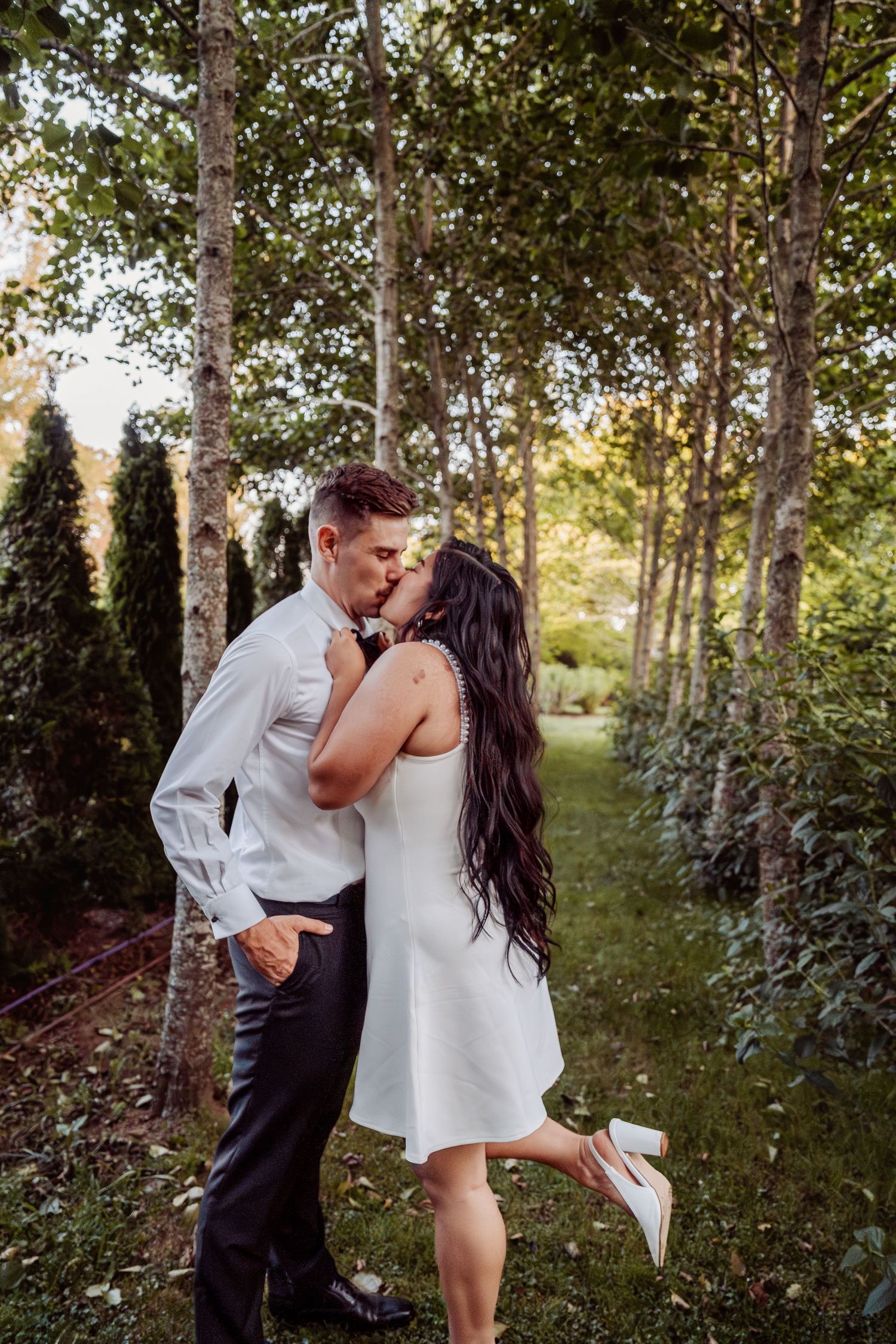 Newlyweds sharing a romantic kiss among tall trees in a sunlit garden path.