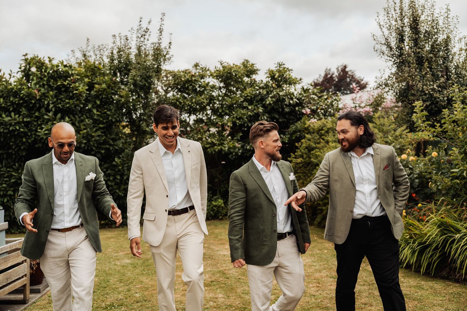 Four groomsmen in green blazers and light suits laughing and walking together outdoors at a wedding.