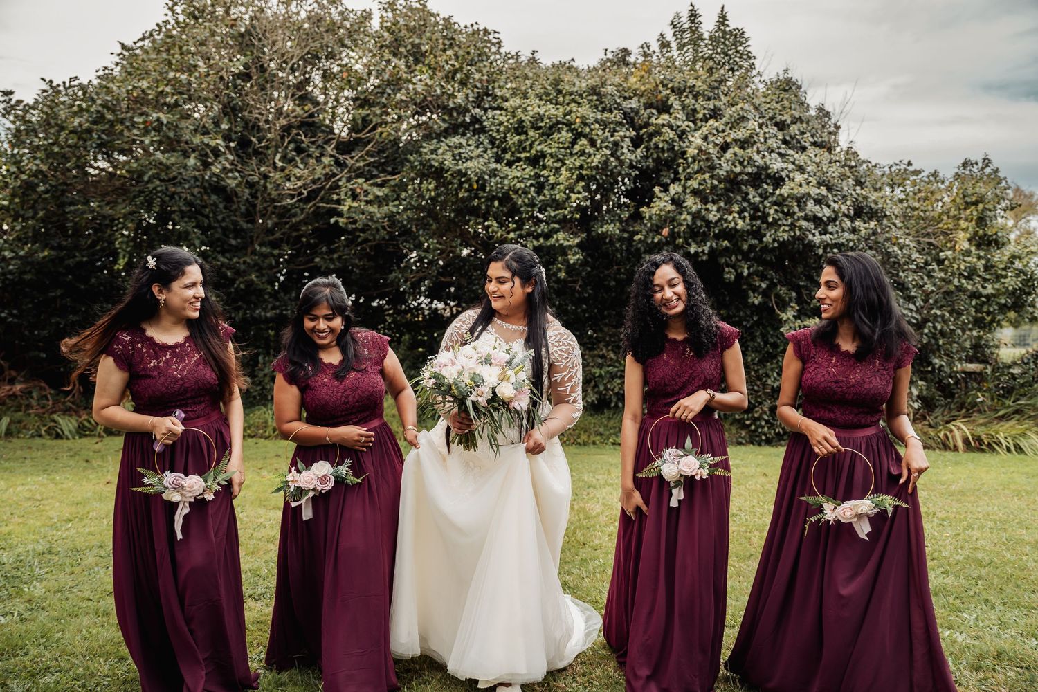 Bride in white gown with four bridesmaids in burgundy dresses holding bouquets on green lawn.