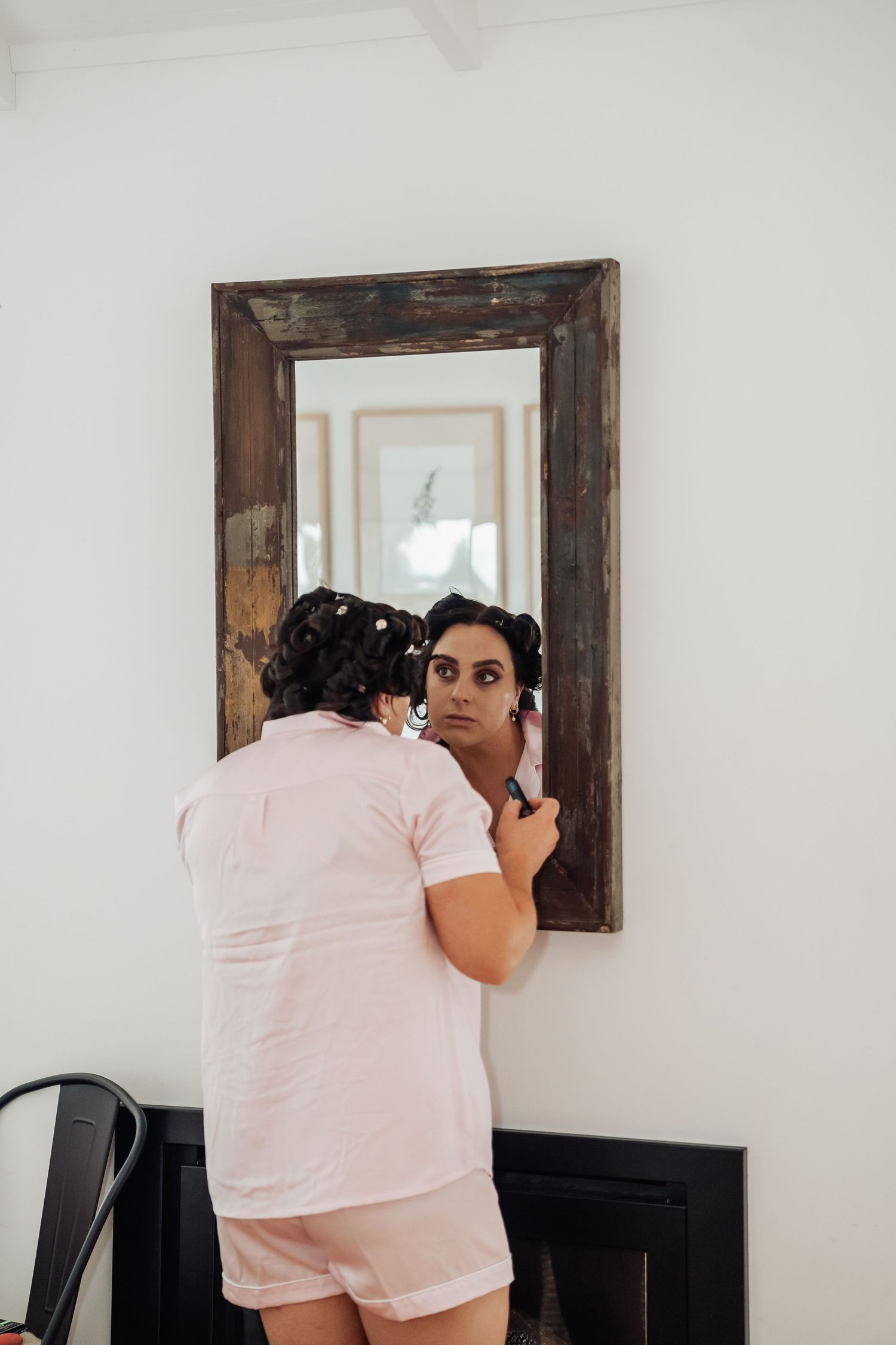Bride in pink pajamas applying makeup while looking into a rustic wooden-framed mirror.