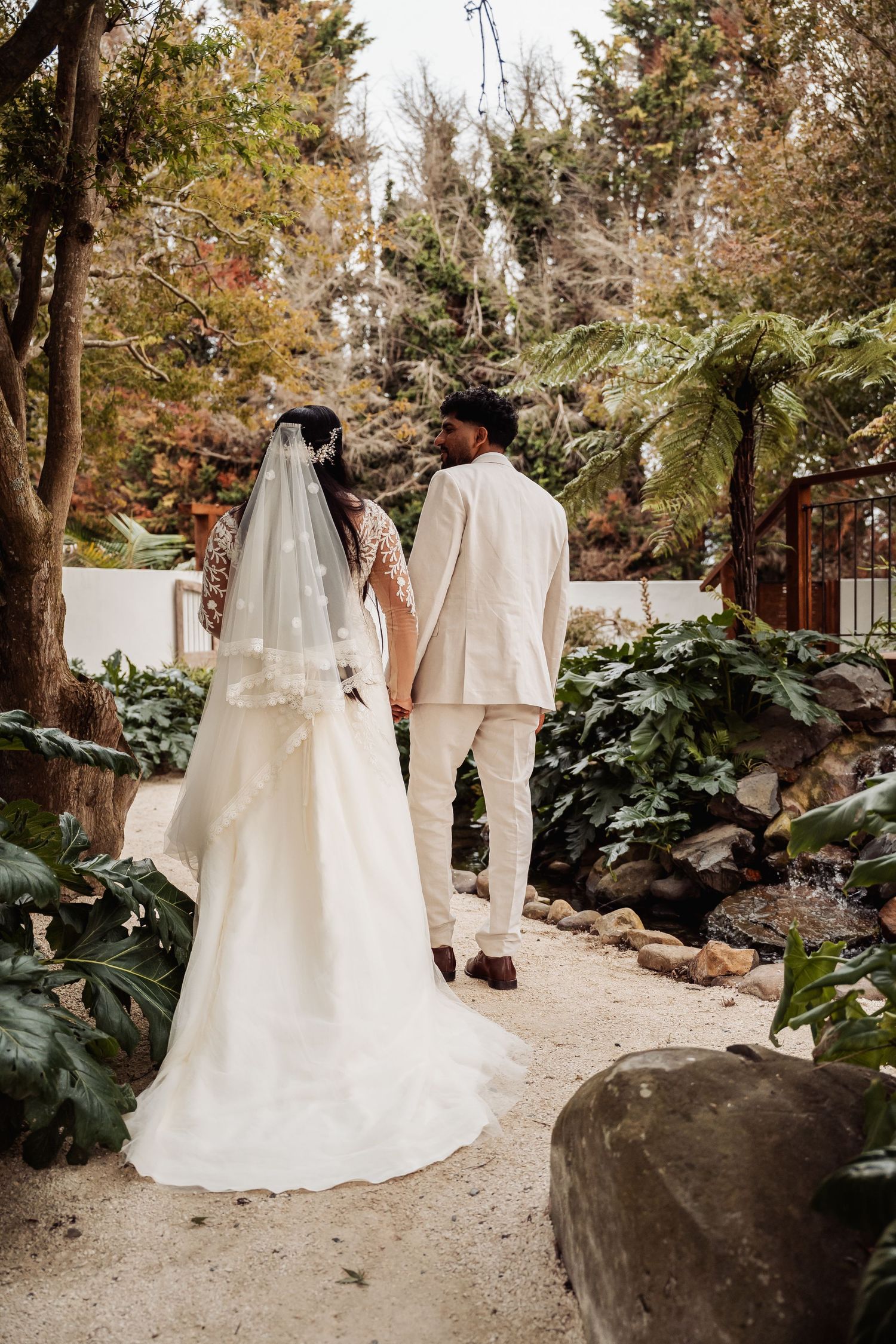 Bride in white gown with veil and groom in cream suit walking hand in hand through garden path.