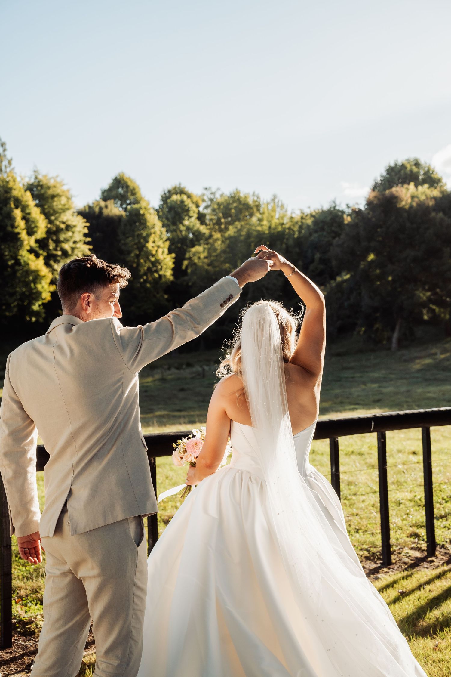 Bride and groom dancing outdoors with raised hands against a scenic green landscape backdrop.